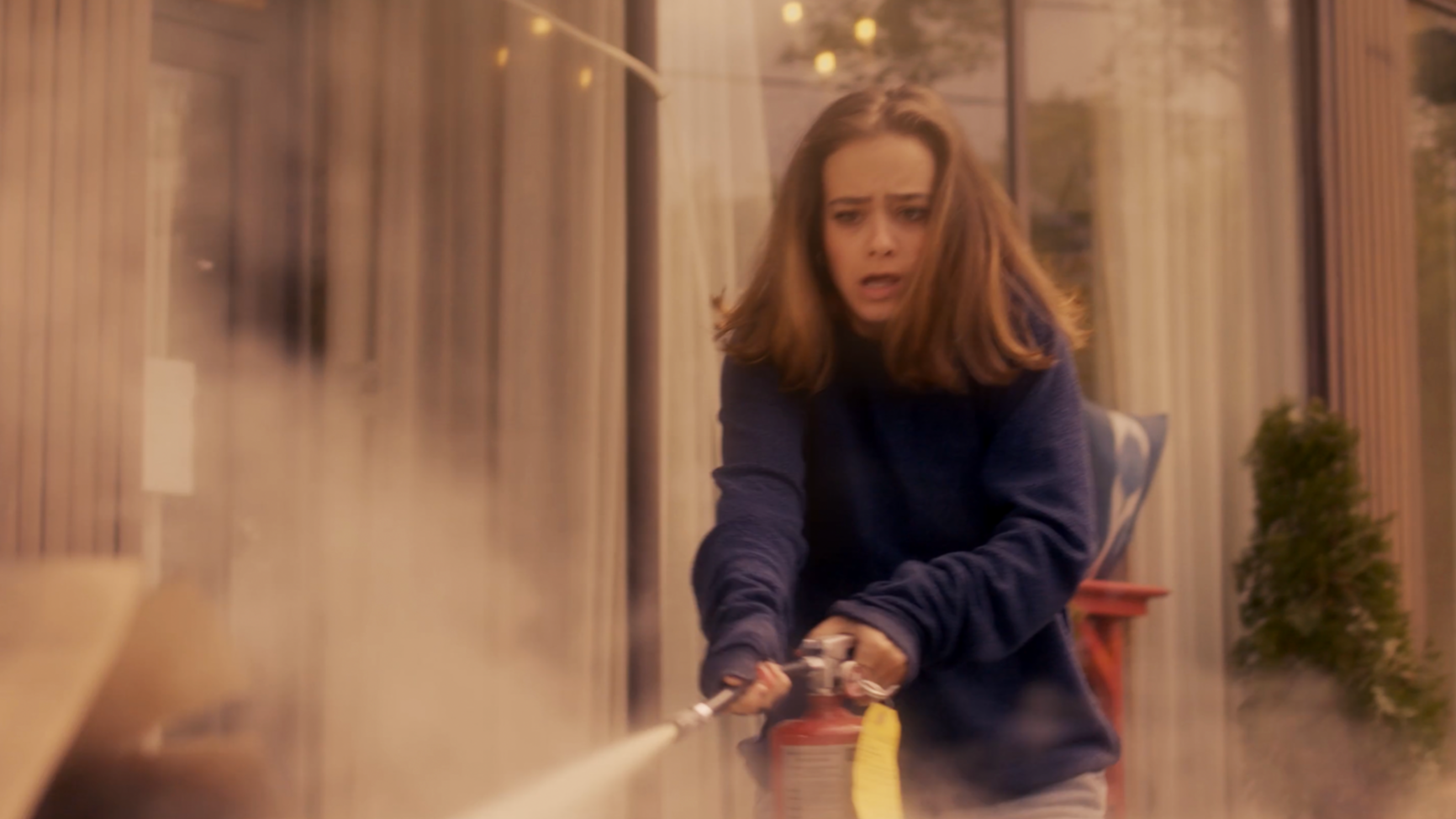 A woman with brown hair wearing a dark blue sweater, holding a fire extinguisher, aiming it at a fire inside a building, with smoke and steam visible.