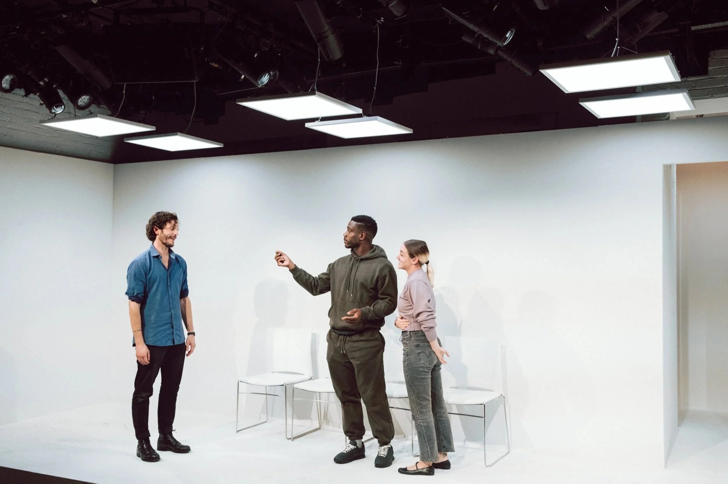 Four people standing and chatting in a minimalist room with white walls and black ceiling panels. The ceiling has several square bright lights. Two chairs are on the floor behind them.