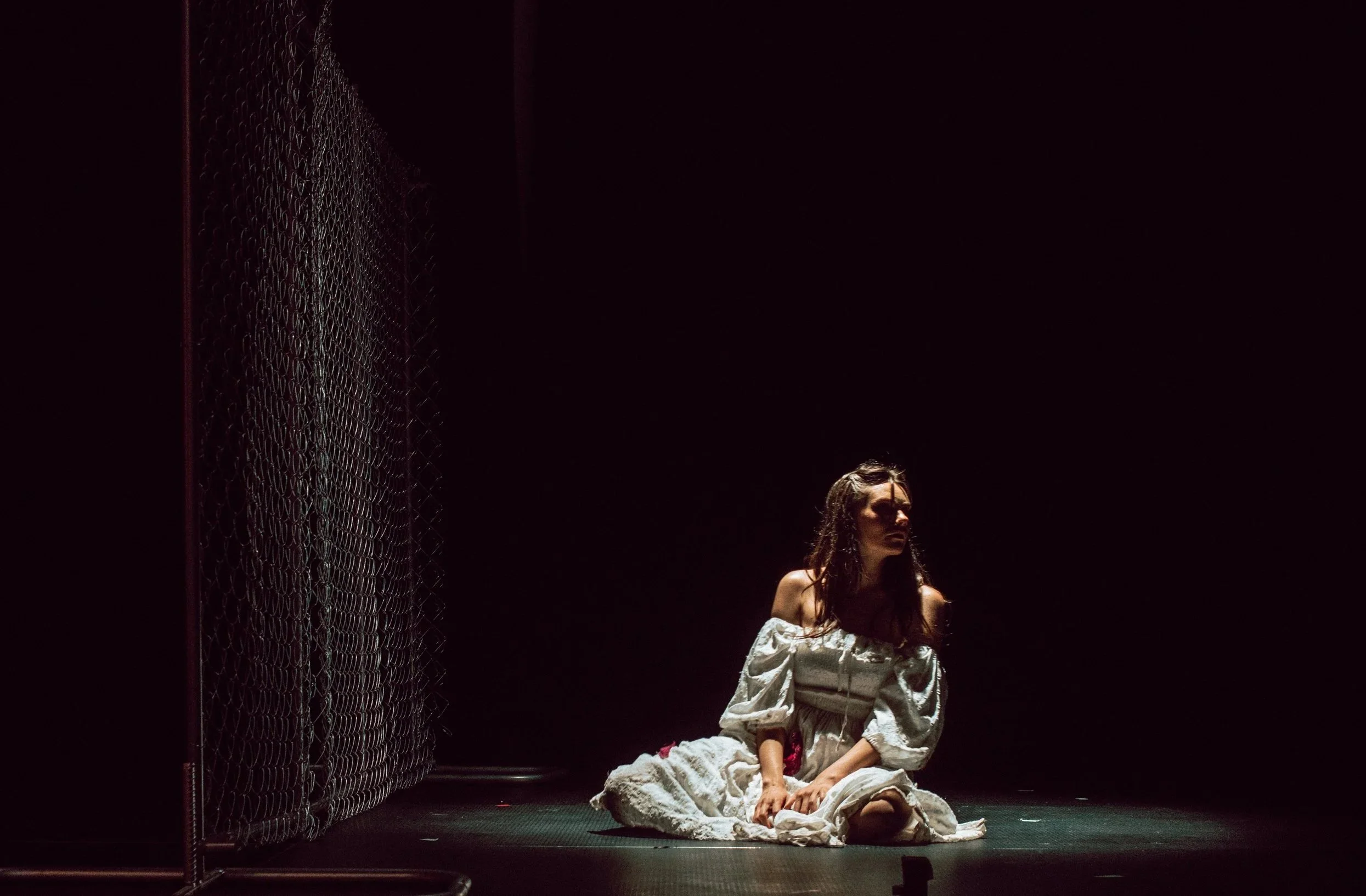 A woman with long dark hair, wearing a white off-shoulder dress, sitting on the ground next to a chain-link fence in a dimly lit space.