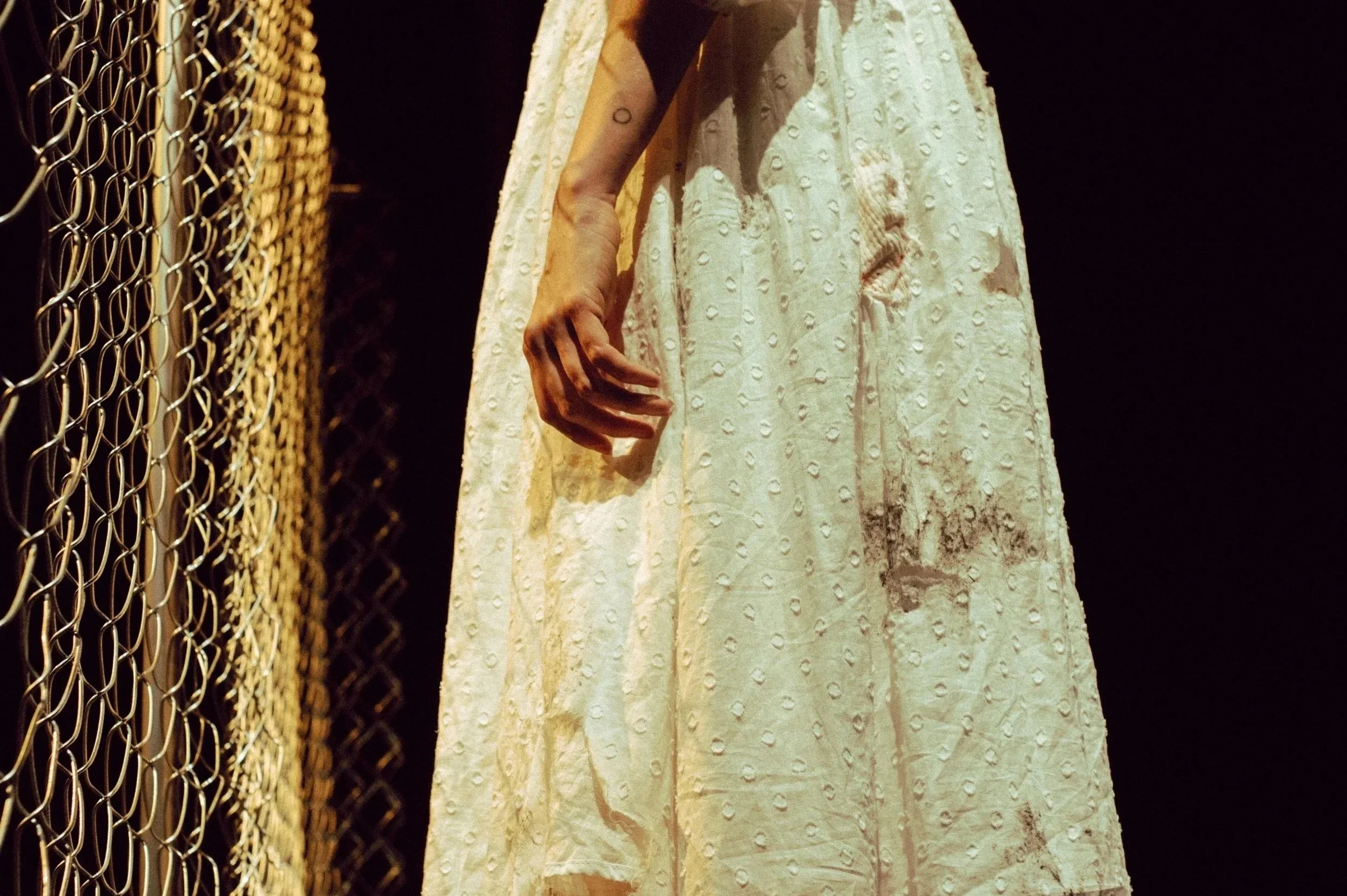 A person wearing a long, white, textured dress with circular patterns, standing next to a metal chain-link fence inside a dimly lit space.