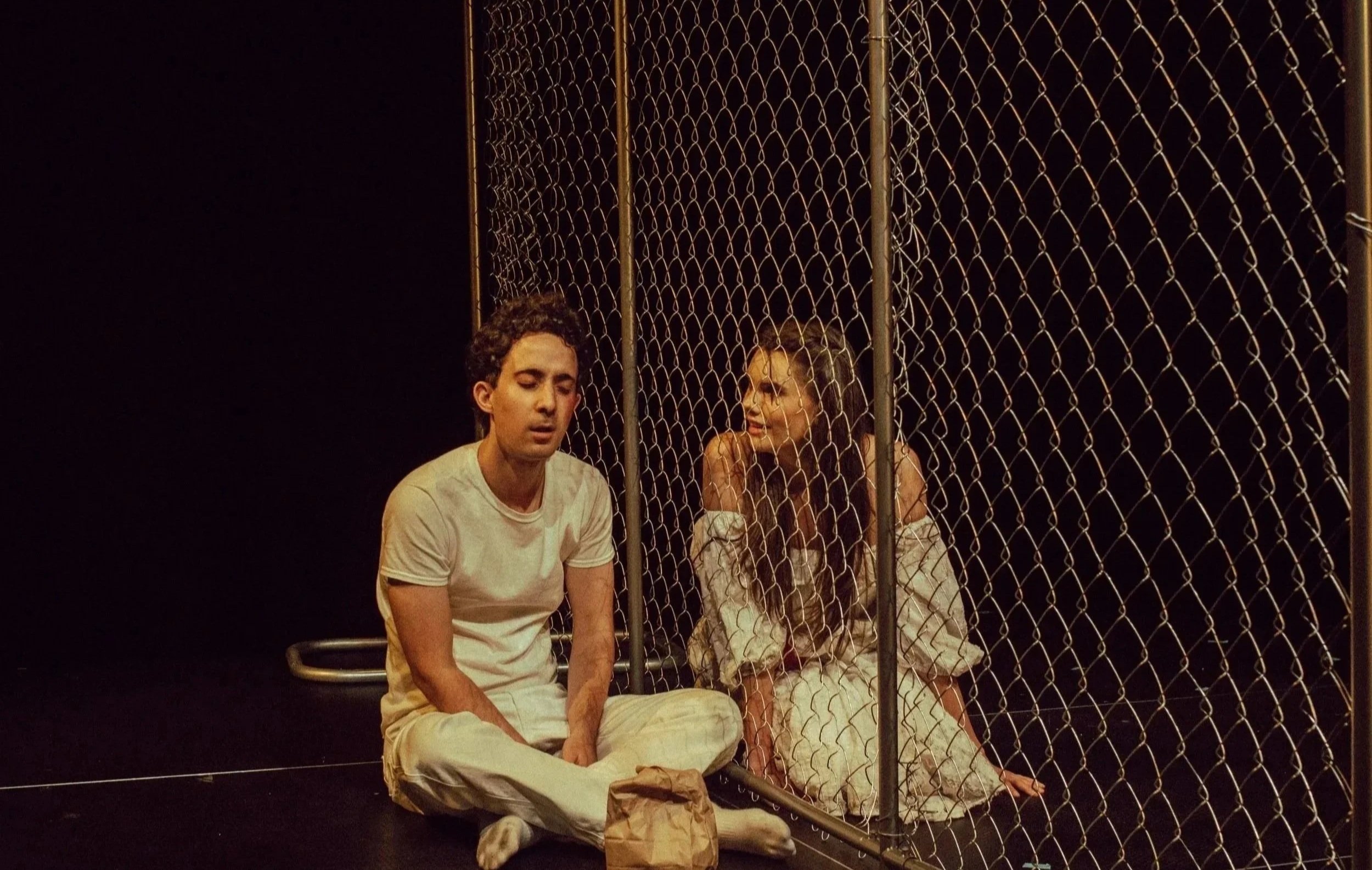 Two people sitting behind a chain-link fence on a dark stage, with a black background. The man on the left has curly dark hair and is wearing a white T-shirt. The woman on the right has long curly hair and is wearing a white jacket. There is a paper 