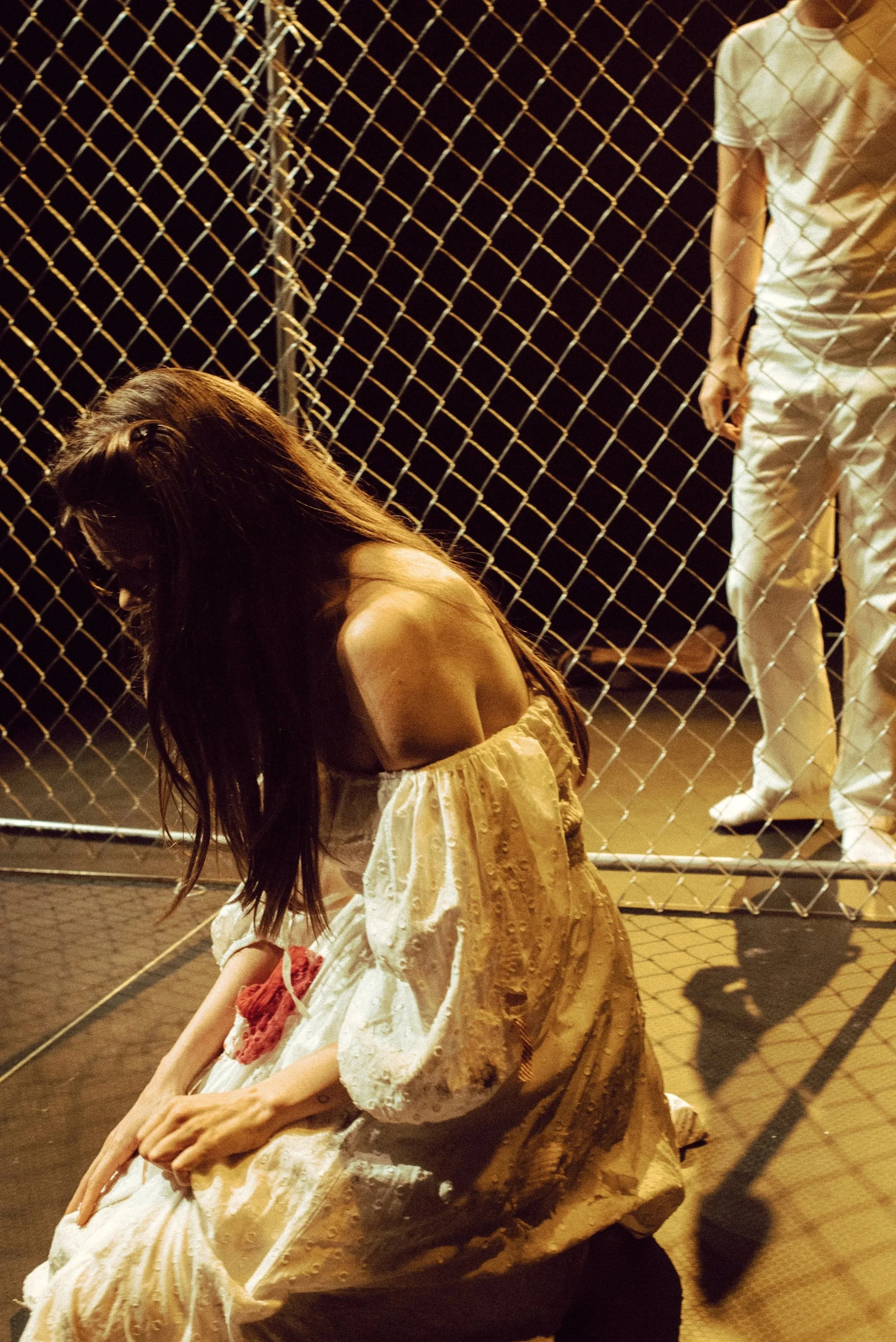 A woman in a white lace dress sitting on the ground at night, with another person standing behind a chain-link fence in a white shirt and pants in the background.