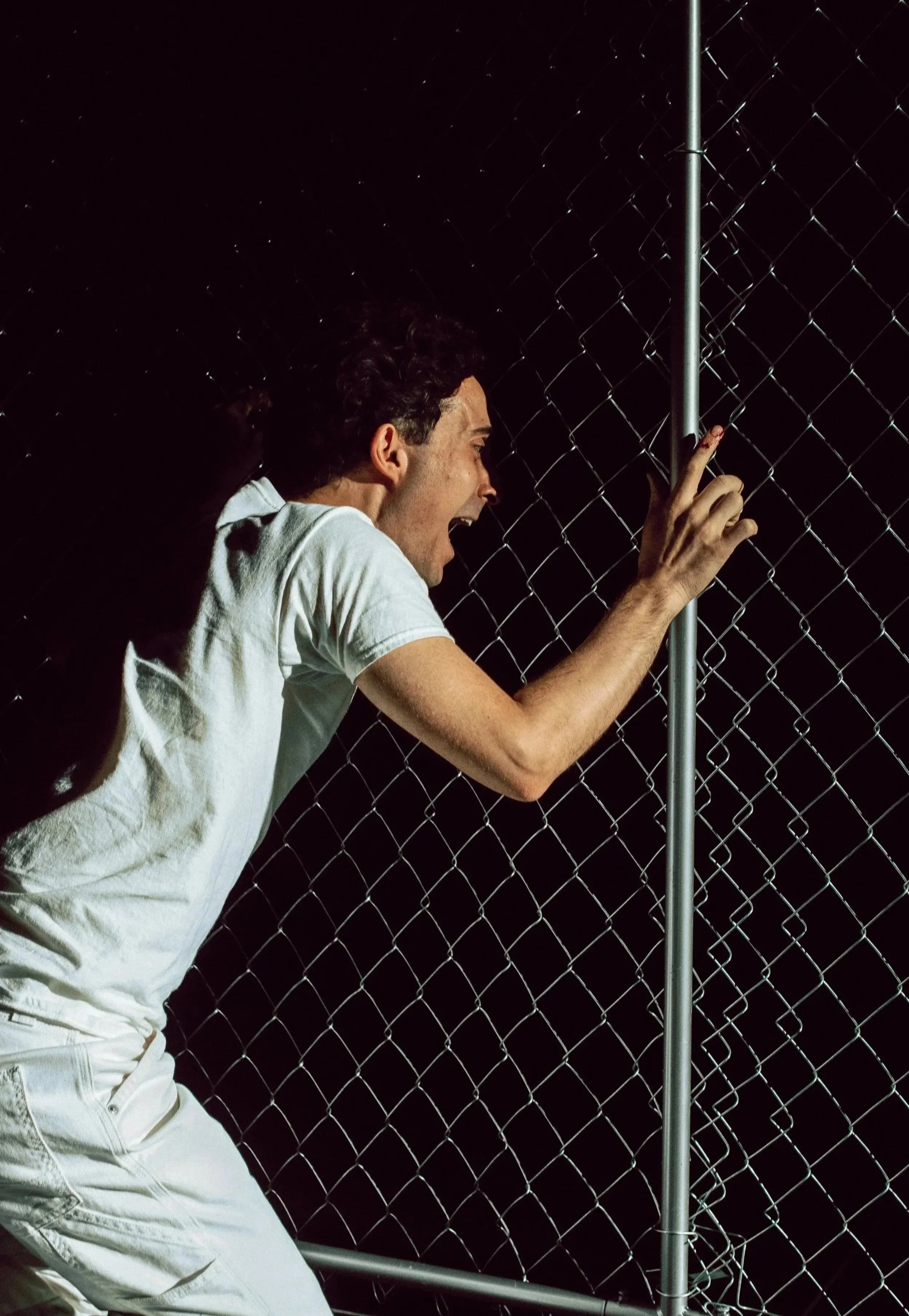 A person with curly dark hair, dressed in a white shirt and white pants, is leaning against a chain-link fence at night, yelling or shouting with their mouth open, facing the fence and holding onto the vertical metal post.