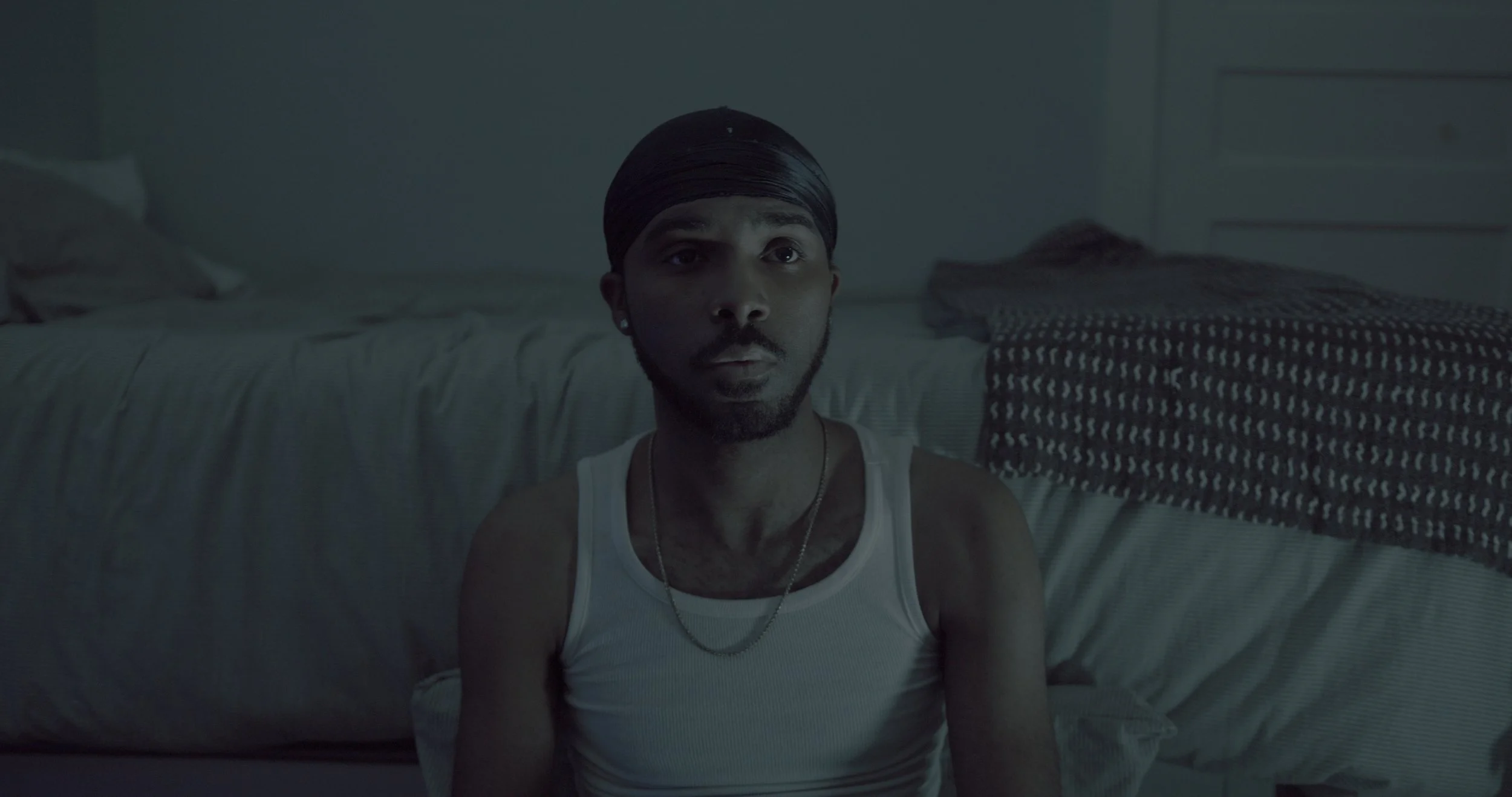 A young man with a beard and mustache, wearing a black durag, silver earrings, a white tank top, and a gold chain, sitting on the floor in a dimly lit bedroom with a bed behind him.