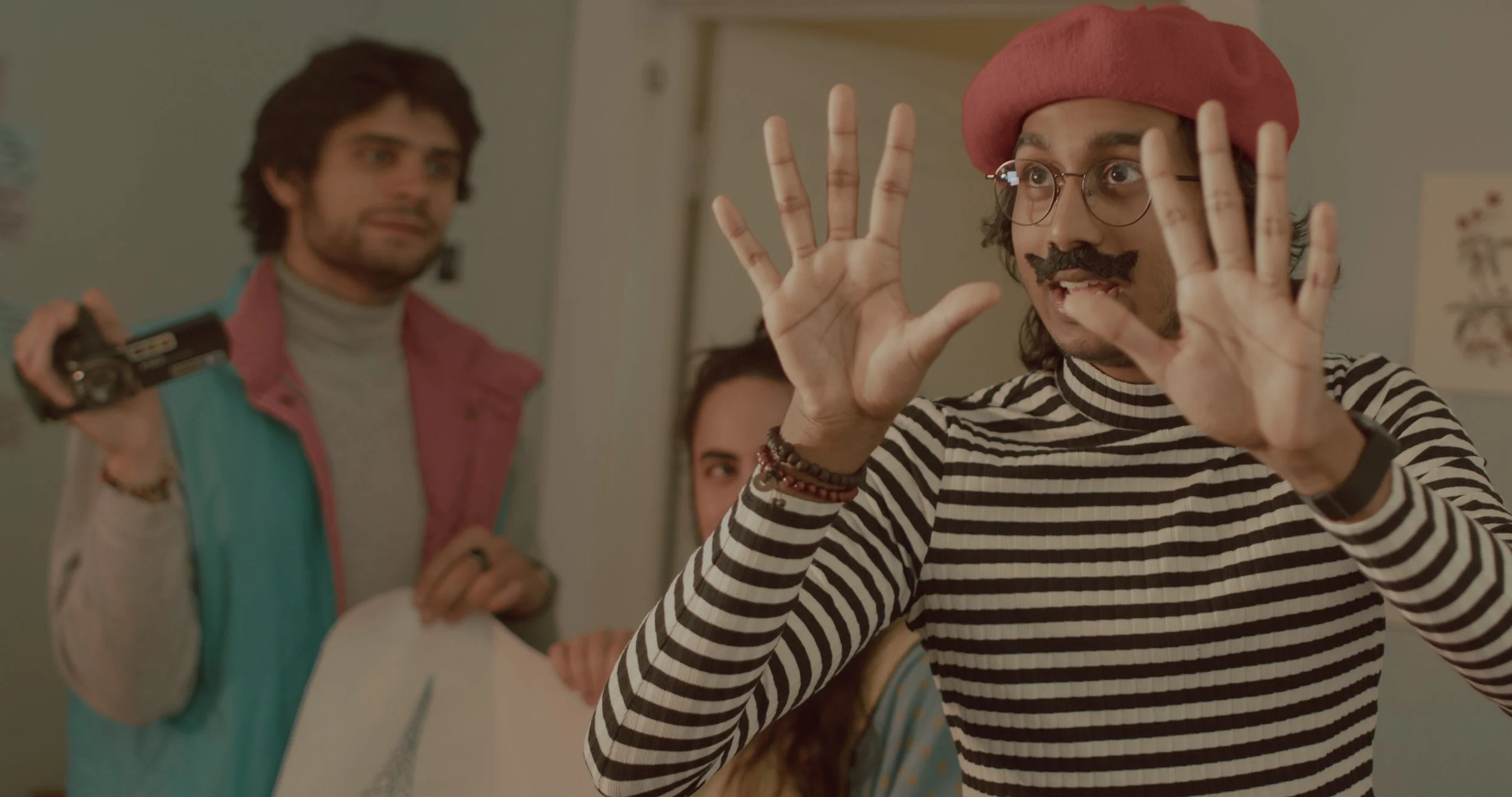 A person wearing a red beret, glasses, a striped shirt, and fake mustache, gesturing with hands. Two others stand behind, one holding a camera, in an indoor setting.