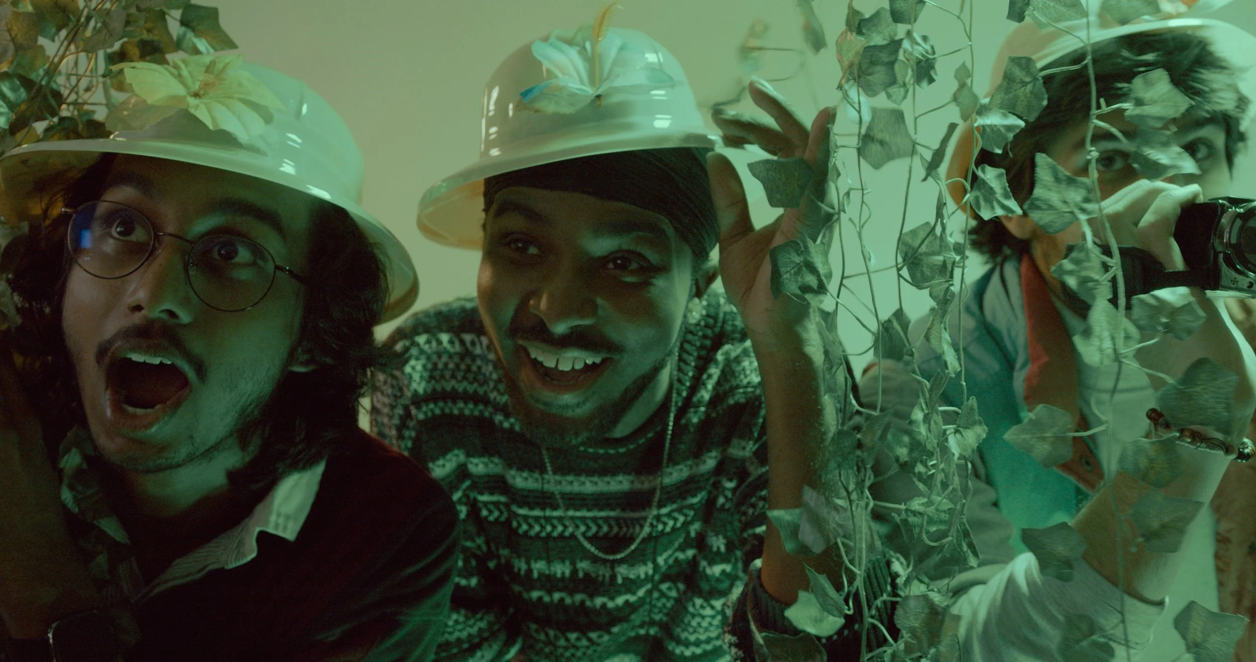 Three people are surrounded by green climbing plants and leaves; they are wearing white hats and are smiling or reacting with surprise.