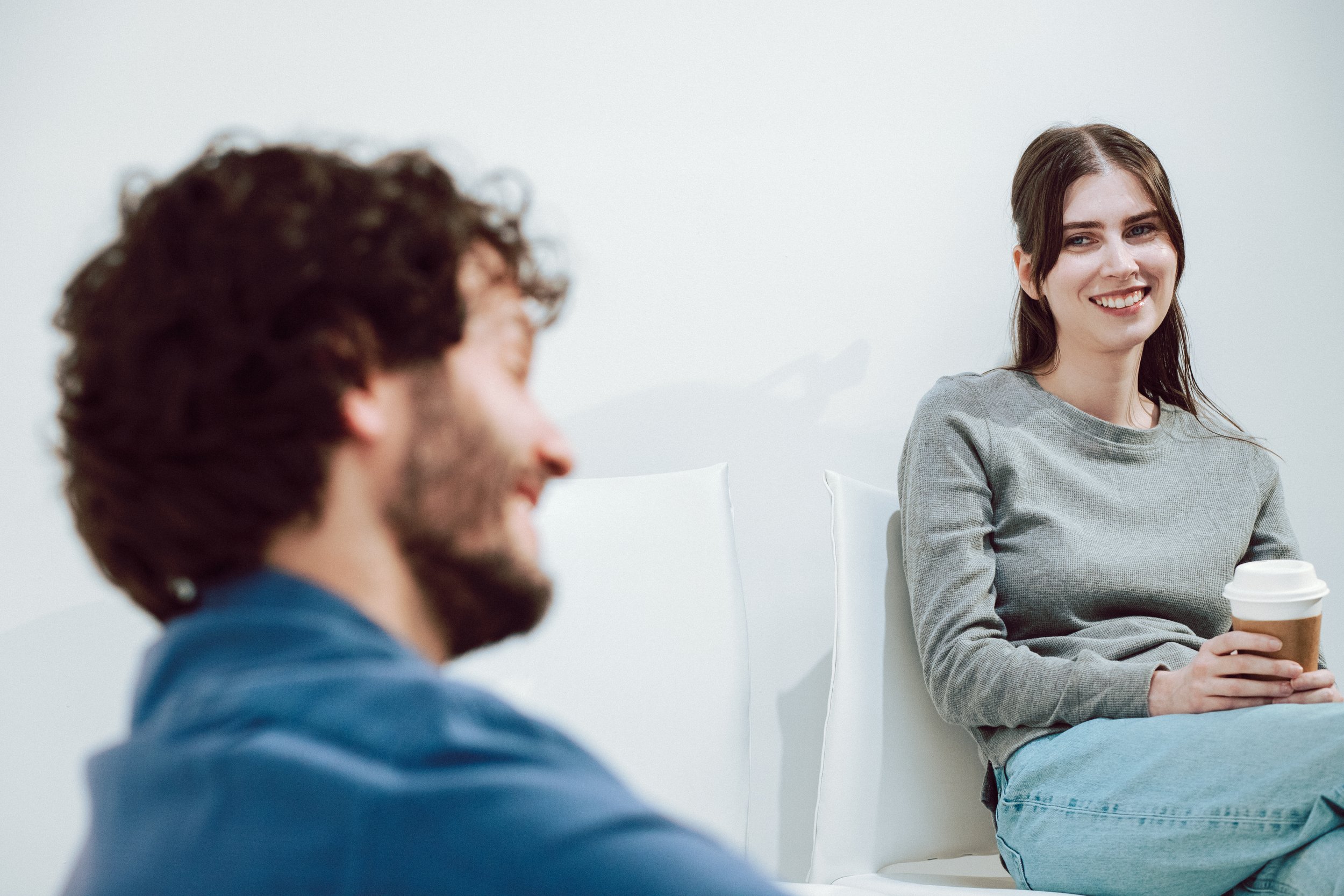 A woman with long dark hair smiling and holding a coffee cup, sitting in a room with plain white walls, while two men are blurred in the foreground, engaging in conversation.