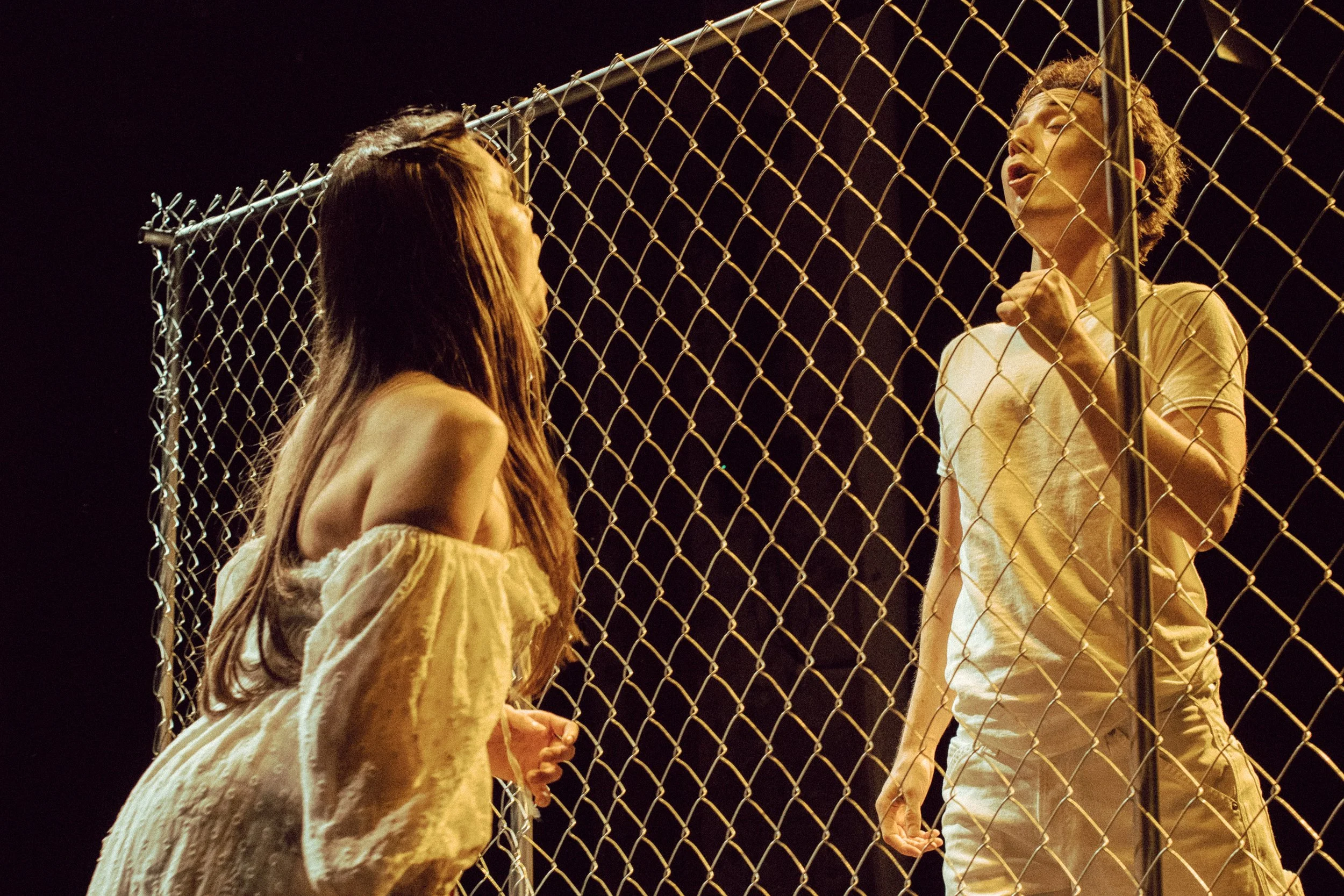 Two women facing each other on either side of a chain-link fence at night, engaging in a serious conversation, with dark background.