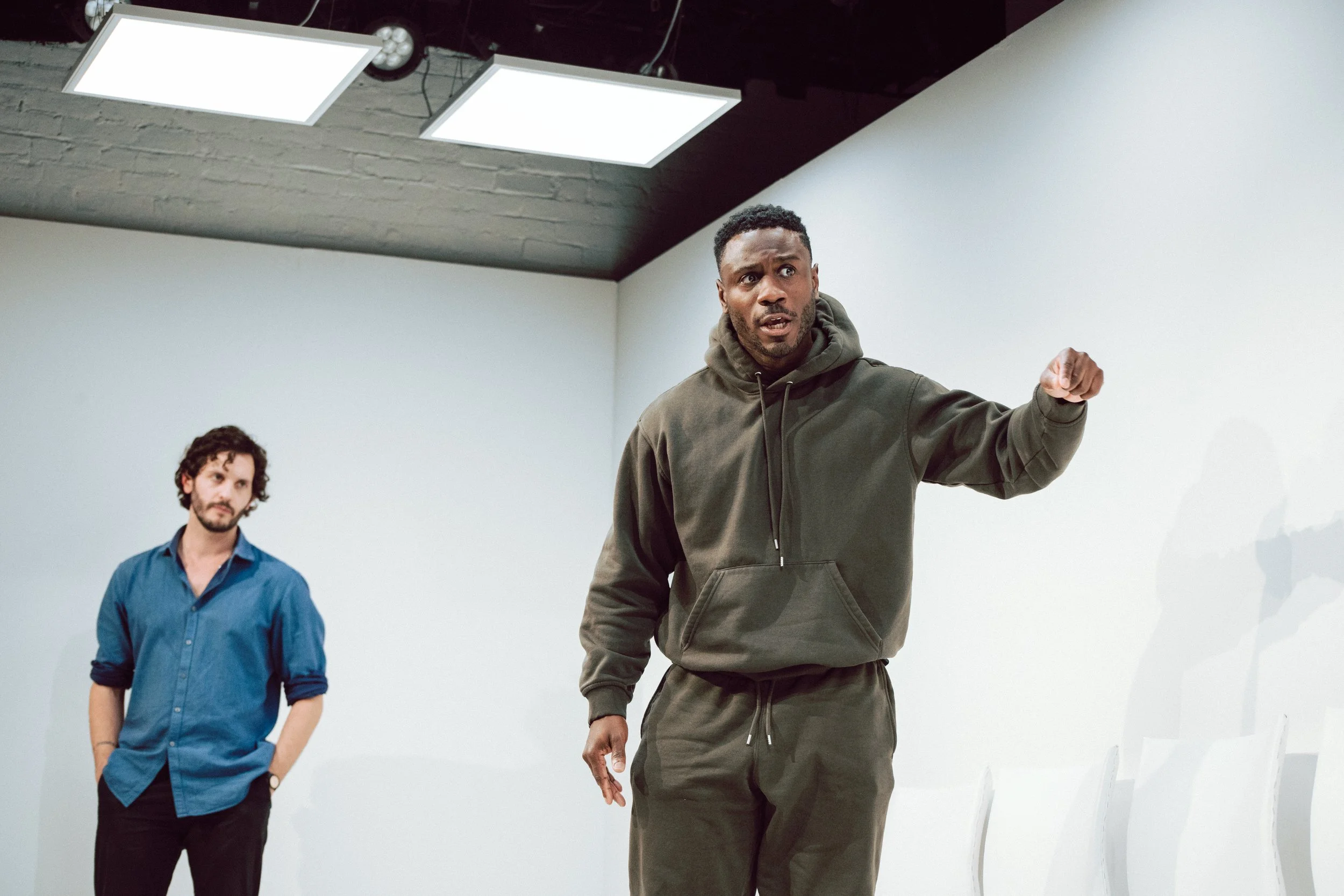 Two men are in a room with white walls. One man is standing and speaking or explaining something, wearing a dark hoodie and pants. The other man, standing slightly behind and to the side, is looking at the speaker with a neutral expression, wearing a