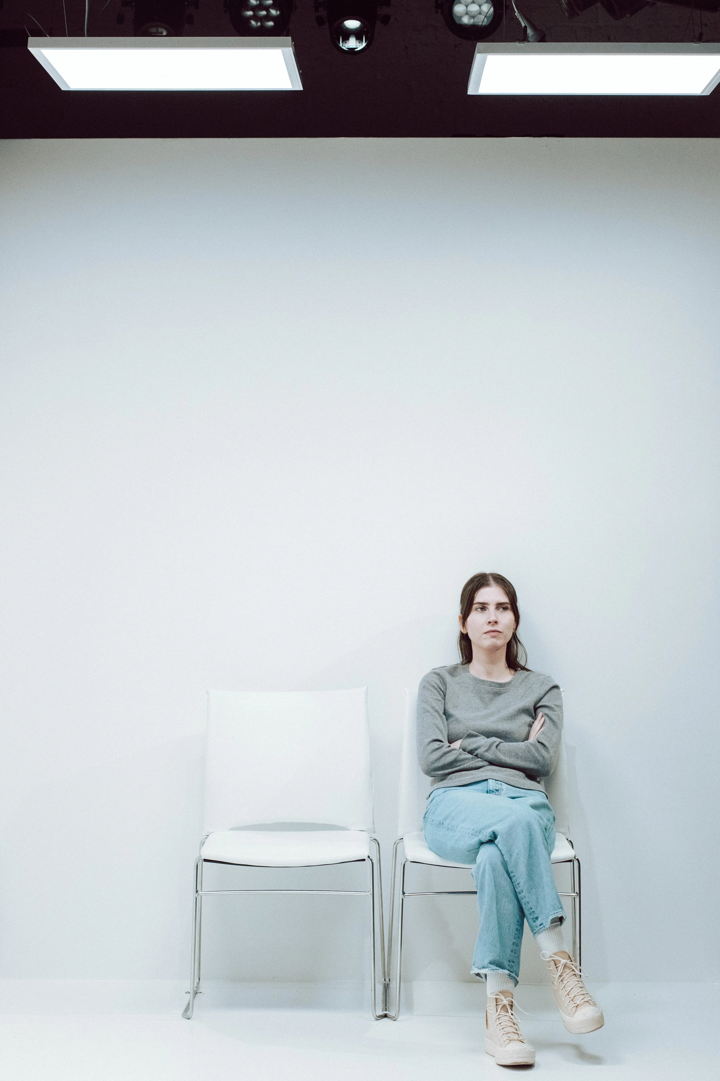 A young woman with long dark hair sitting on a white chair in an empty white room with two chairs and a plain white wall, crossing her arms and wearing a grey sweatshirt, light blue jeans, and beige sneakers.