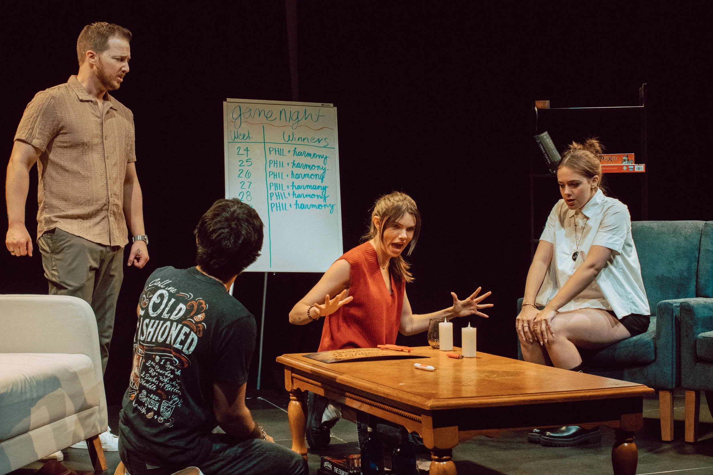 People gathered in a living room playing a game. A woman in a red top is talking animatedly, while a man and a woman are sitting and listening. A man in a patterned shirt is standing near a whiteboard with game instructions, and another woman is seat