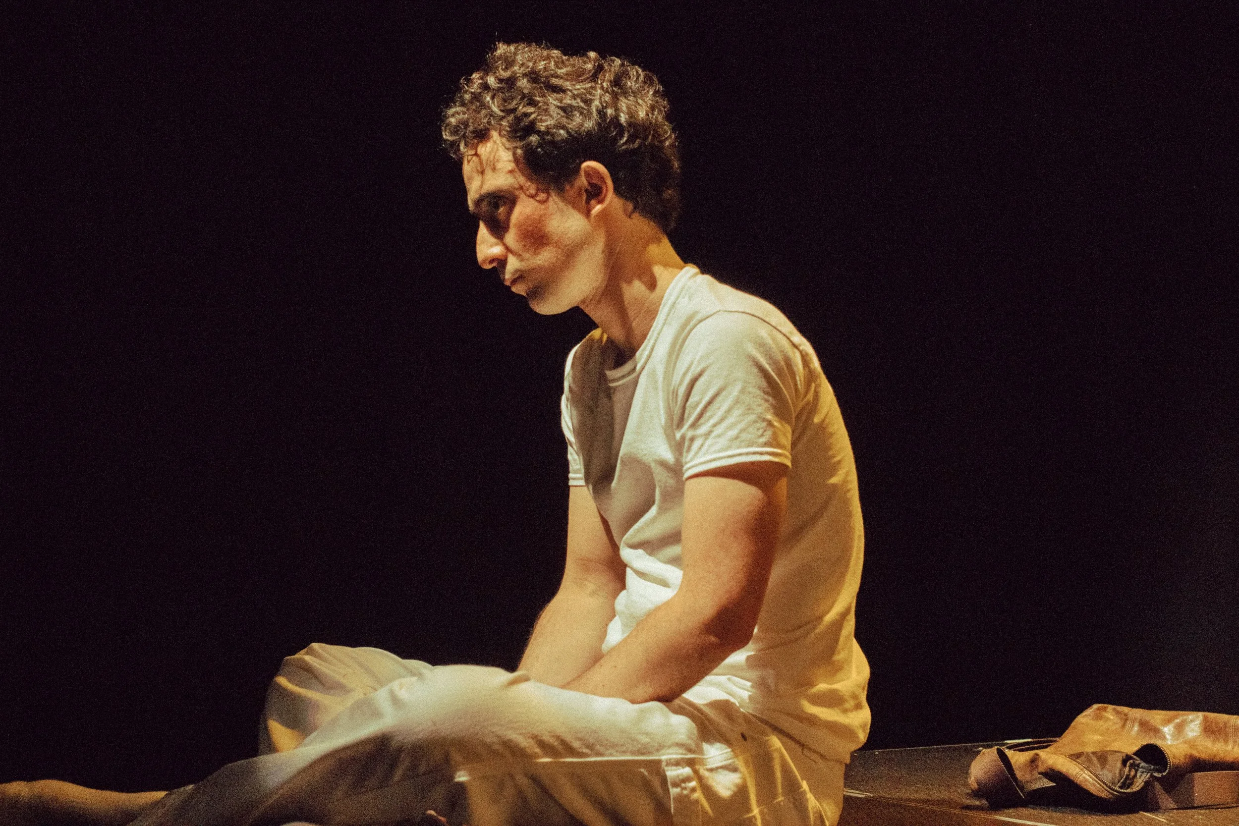 A person with curly hair wearing a white shirt sitting on a stage or platform, looking down with a serious or somber expression, against a dark background.