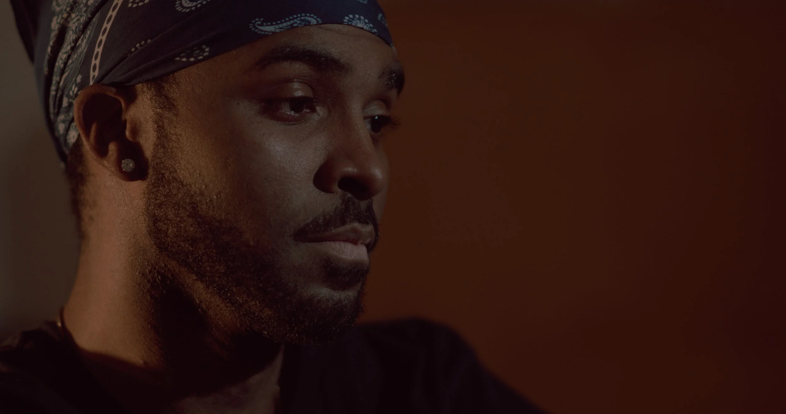 Close-up of a person's face with dark skin, wearing a black bandana with white patterns, looking down with a calm expression.