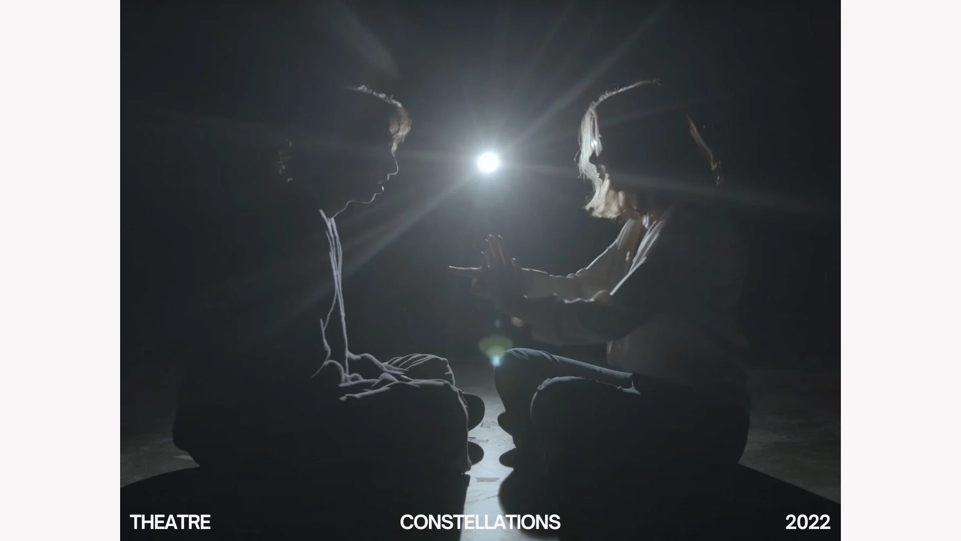 Two people sitting cross-legged facing each other in darkness with a bright light between them, reaching out to touch or hold each other's hands or fingers.