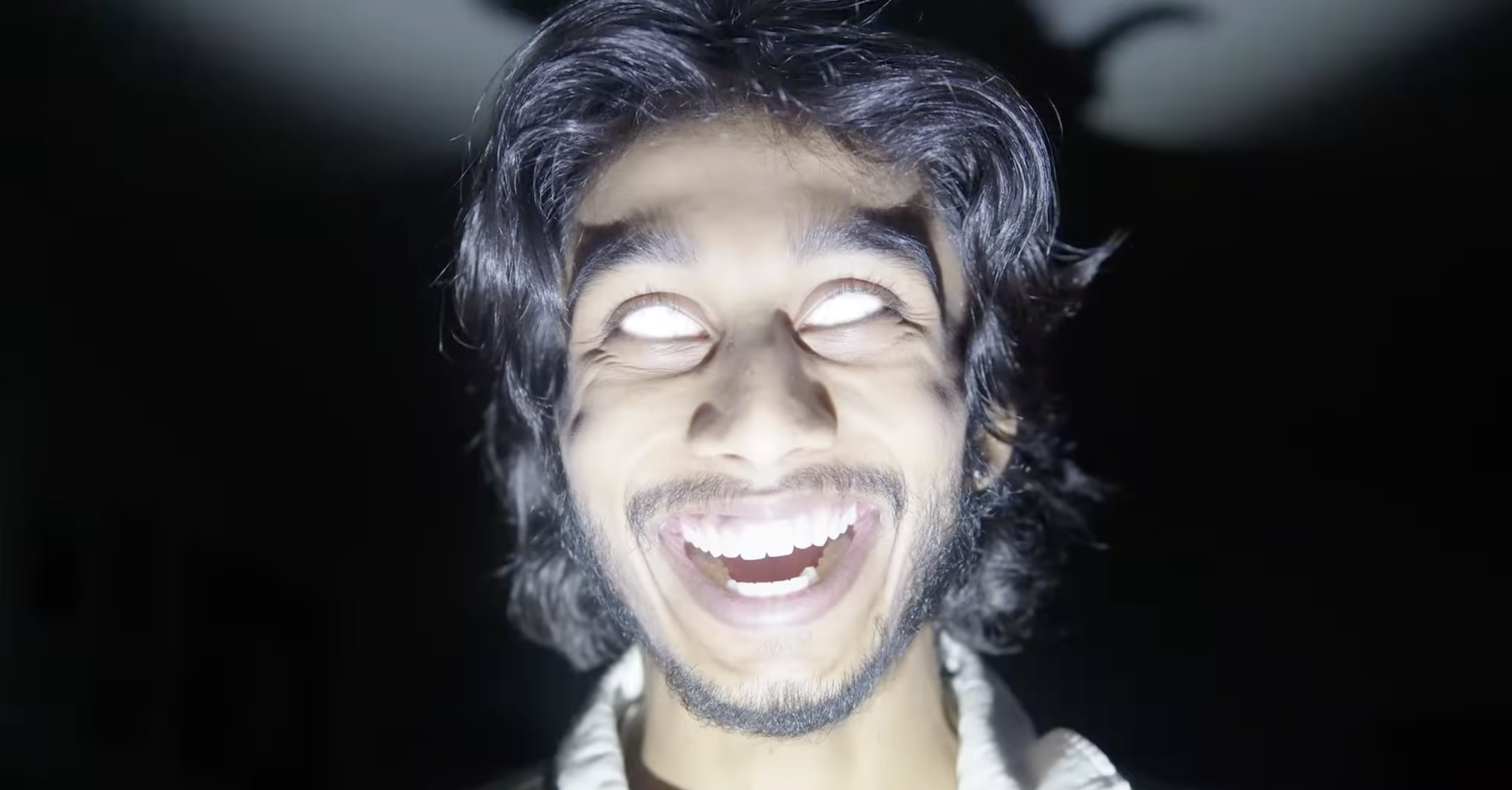A young man with dark, wavy hair and a beard, smiling widely with his teeth visible, against a dark background.