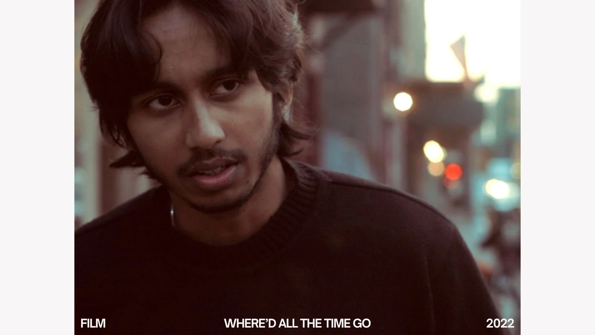 Close-up of a young man with wavy hair and a mustache, wearing a dark sweater, standing outdoors on a city street at dusk with blurred city lights in the background. 