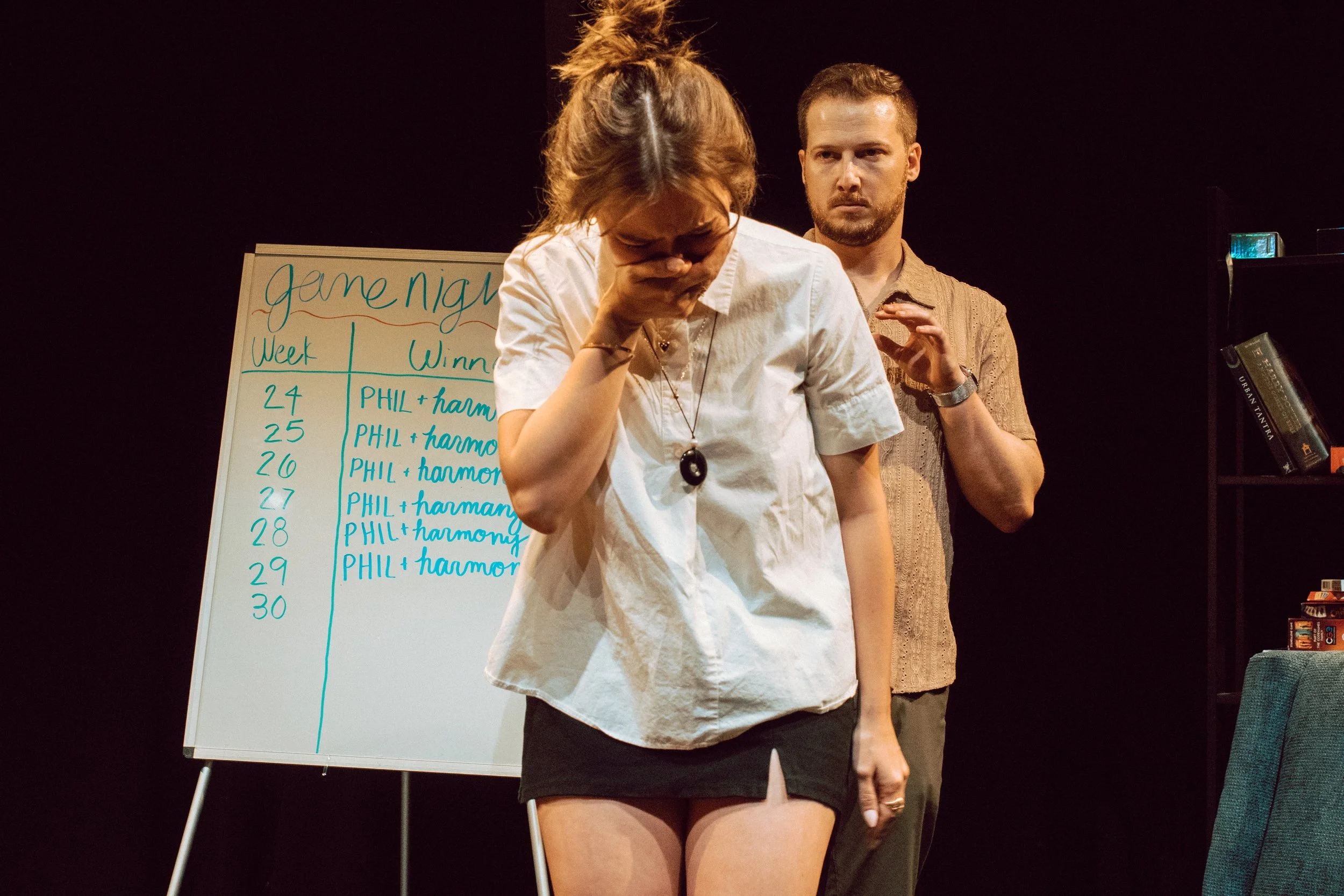 Two people standing in front of a whiteboard, one with curly hair tied up, wearing a white shirt and shorts, holding her head in her hand, and the other with short hair, beard, wearing a brown shirt, looking at her. The whiteboard has a list with dat