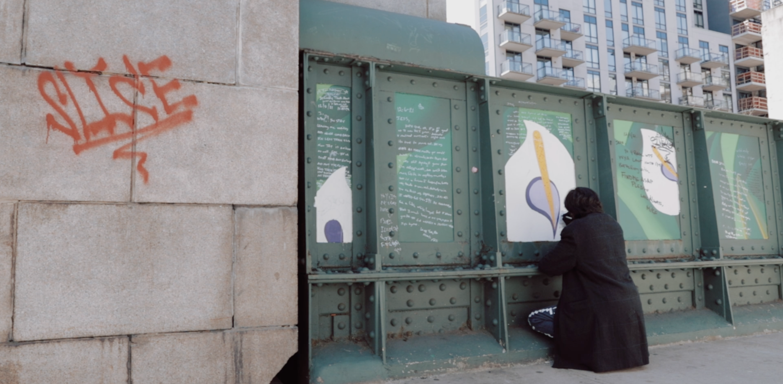 A person kneeling in front of a green metal structure with posters or signs on it, located in an urban setting with high-rise buildings in the background. A red graffiti tag is on the stone wall to the left.