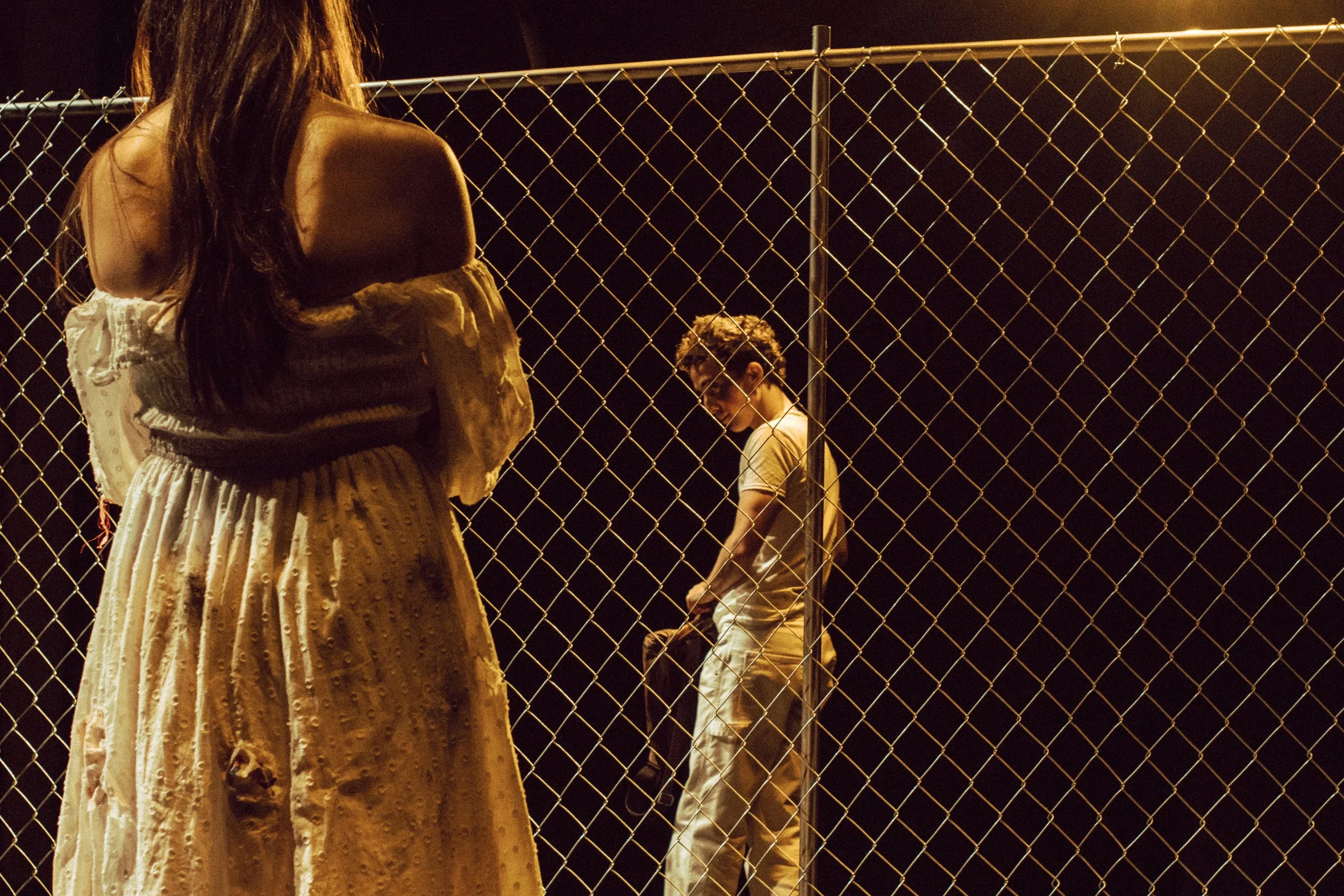 A woman with long dark hair wearing a beige dress standing next to a chain-link fence at night. In the background, a man in a beige jacket and glasses sitting on the ground, looking downward.