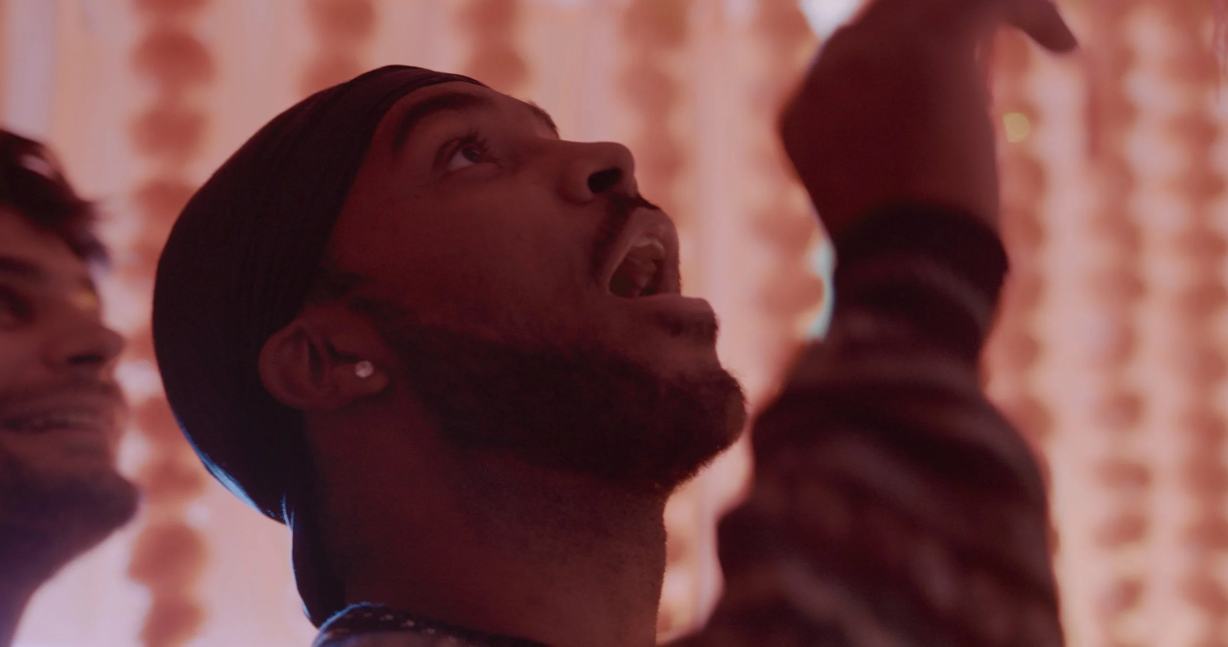 A man with a beard and earring, wearing a headband, looking upwards, with other blurred people's faces around him, in a dimly lit setting with warm, orange lighting.