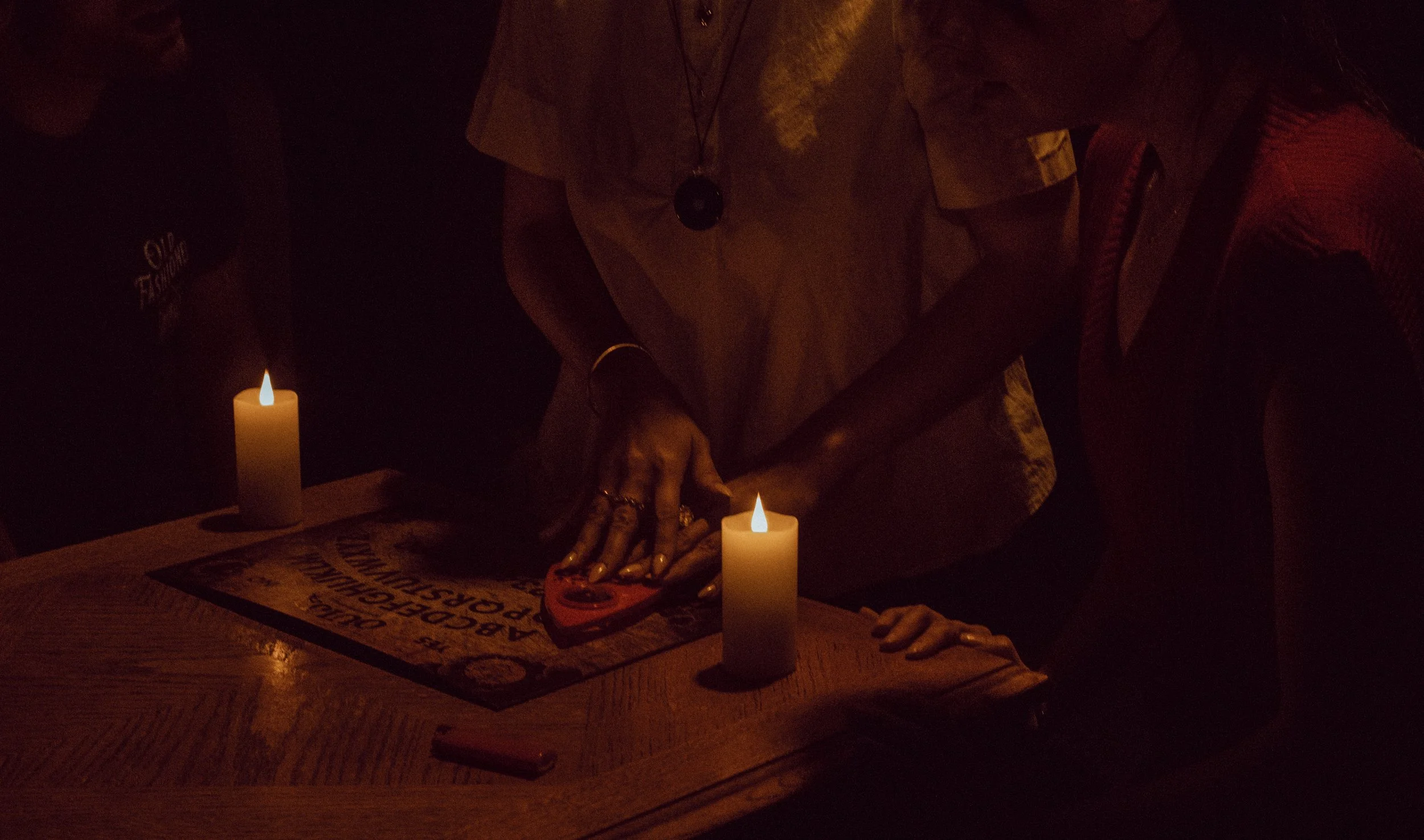 Person holding a planchette over a Ouija board with candles on a table in a dimly lit room.