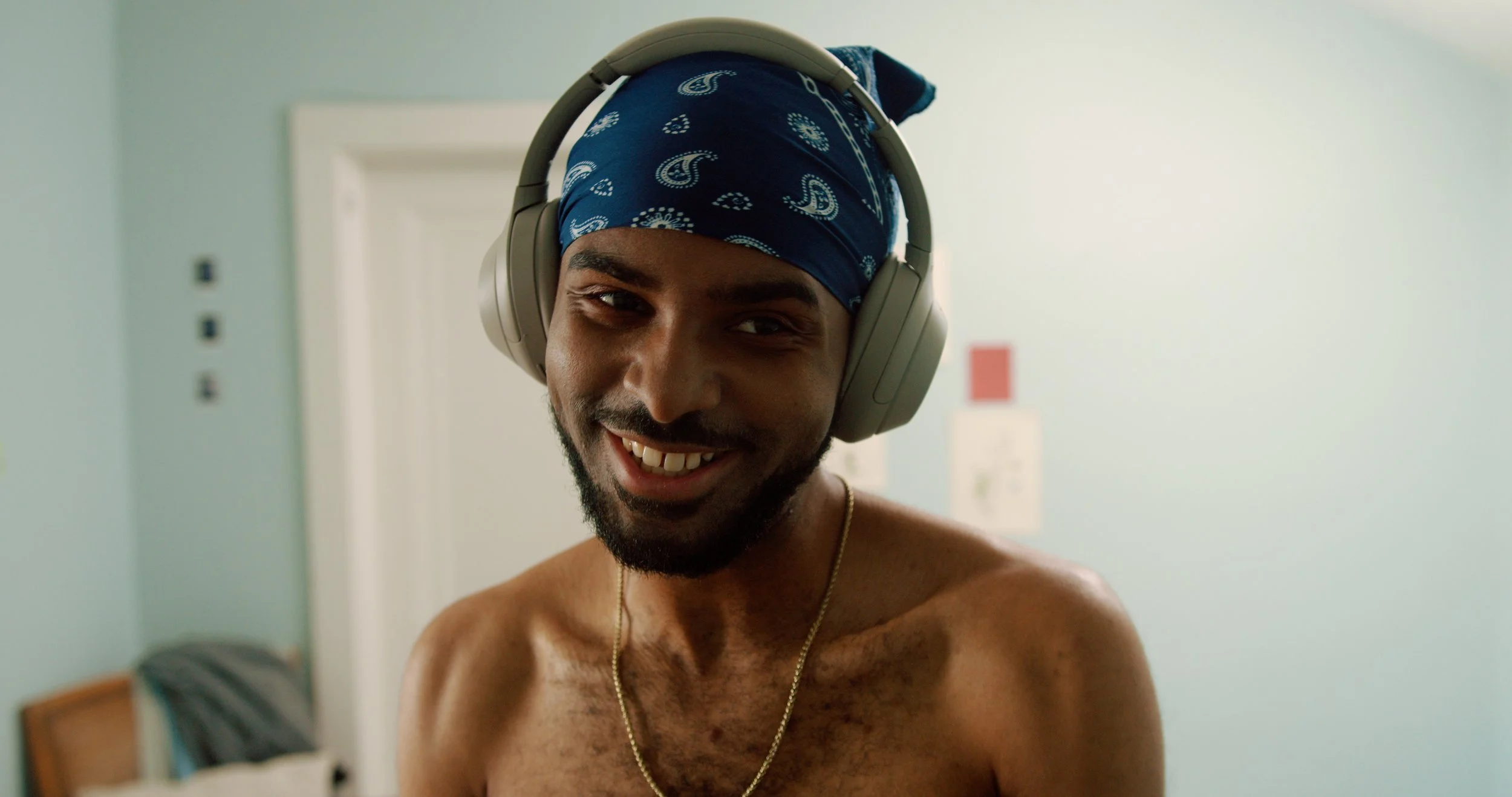 A shirtless man wearing a blue bandana and large over-ear headphones, smiling inside a room with pale green walls.