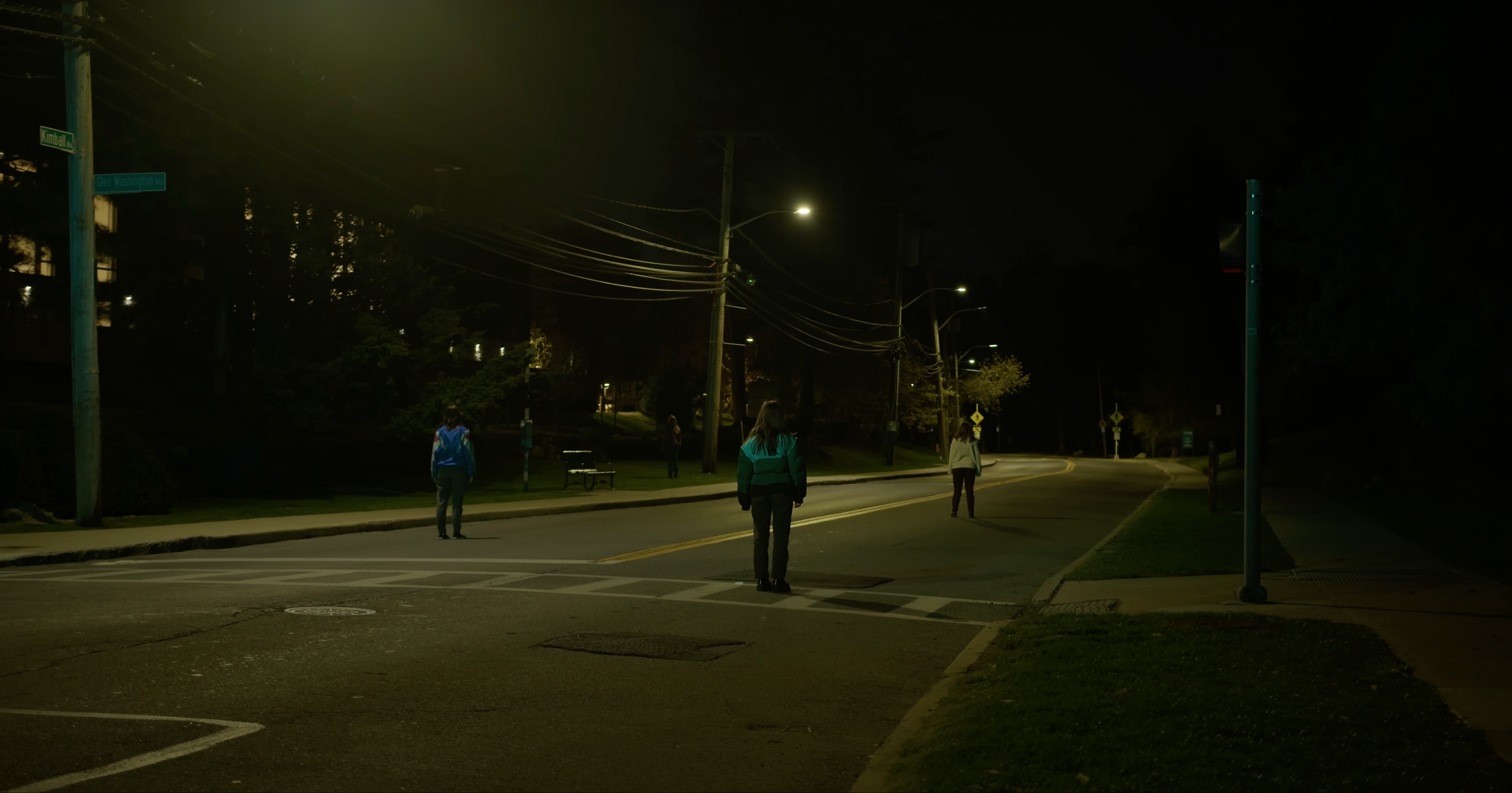 A street at night with three people standing on the crosswalk, illuminated by streetlights with power lines overhead and trees in the background.