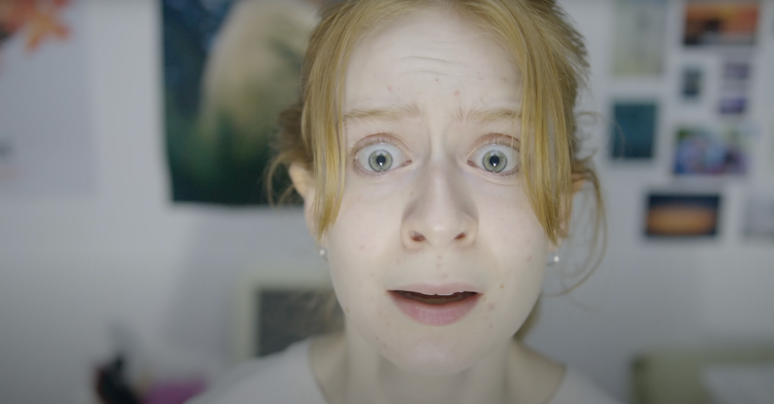 A young woman with red hair and green eyes, looking surprised or amazed, with her mouth slightly open. She has a light complexion with a few blemishes and is in an indoor setting with photo wall decor in the background.