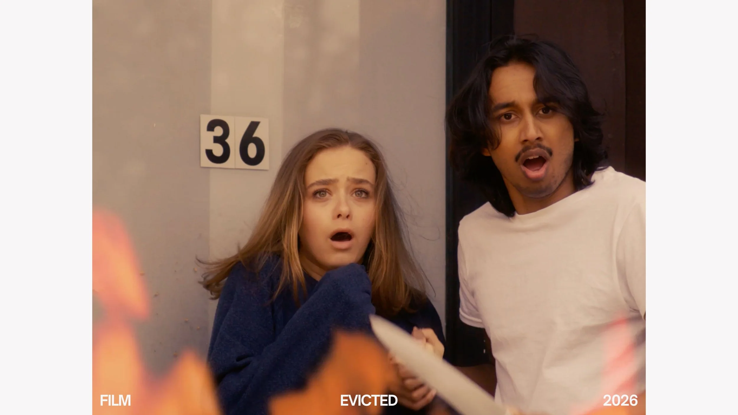 A young woman with long brown hair and a young man with shoulder-length black hair stand side by side, looking surprised or shocked. The woman is wearing a dark blue jacket, and the man is wearing a white t-shirt. There is a wall clock showing the nu