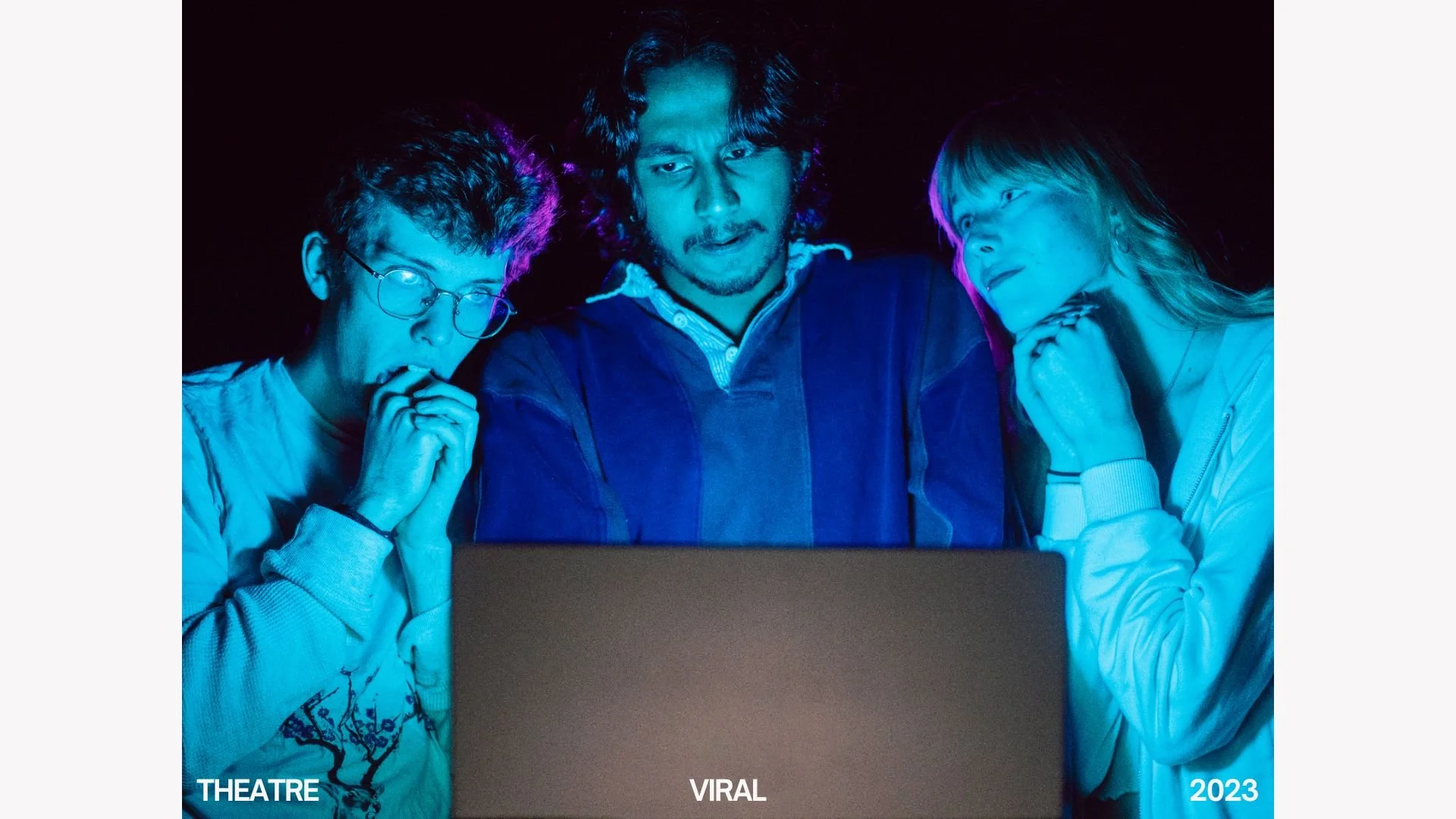 Three people looking at a laptop screen in a dark room, illuminated by blue light. The person in the middle has curly hair and a beard, wearing a dark jacket. The person on the left has glasses and curly hair, wearing a light-colored sweatshirt. The 