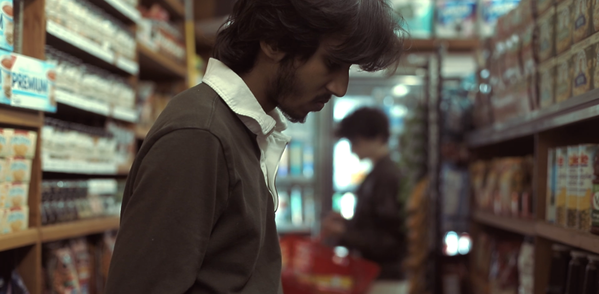 A man with brown hair and a beard shopping in a grocery store aisle, looking at products on the shelves.