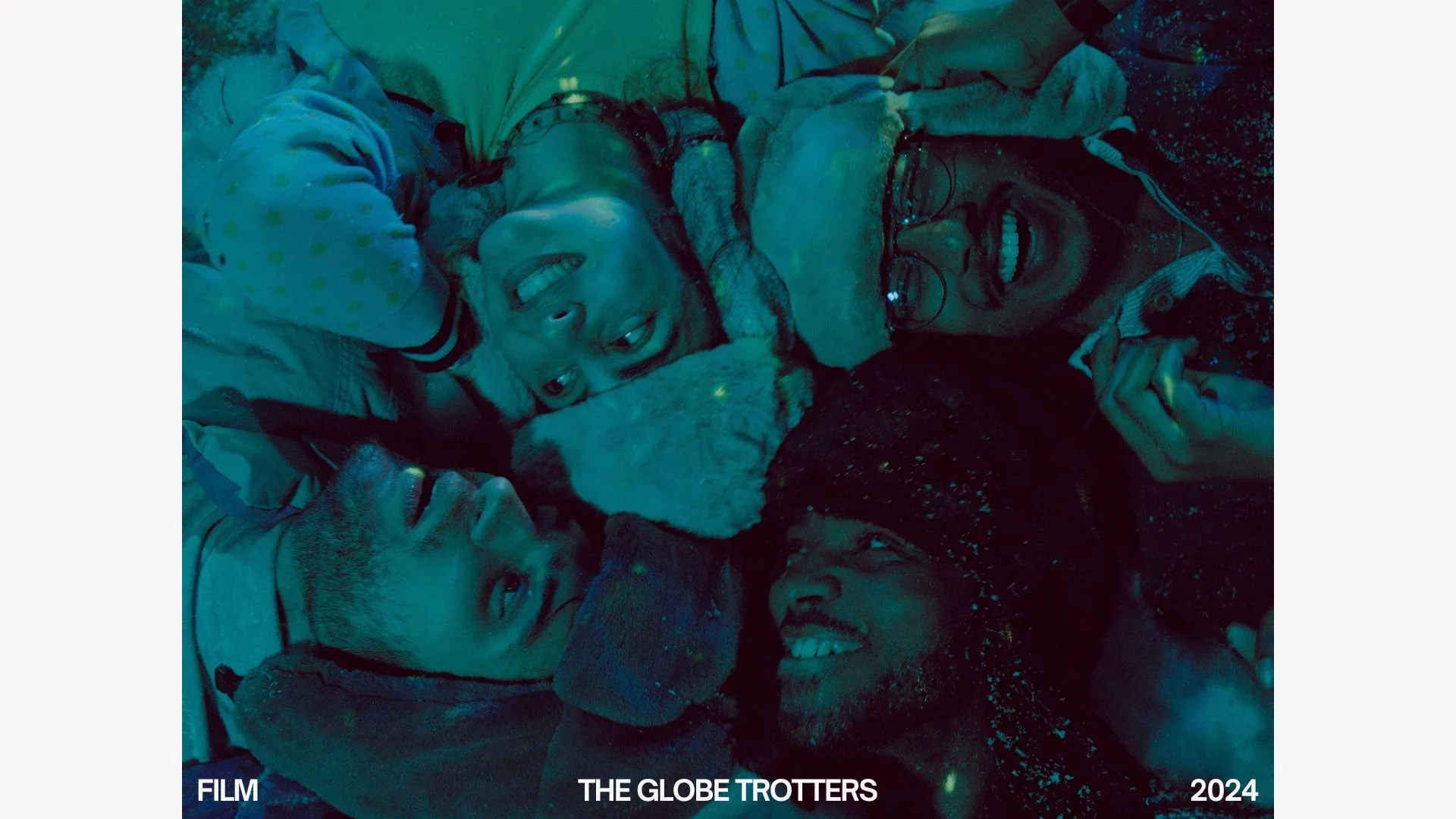 A group of young people lying on the ground together, smiling and cuddling with a dog, illuminated by blue-green light in a nighttime setting.