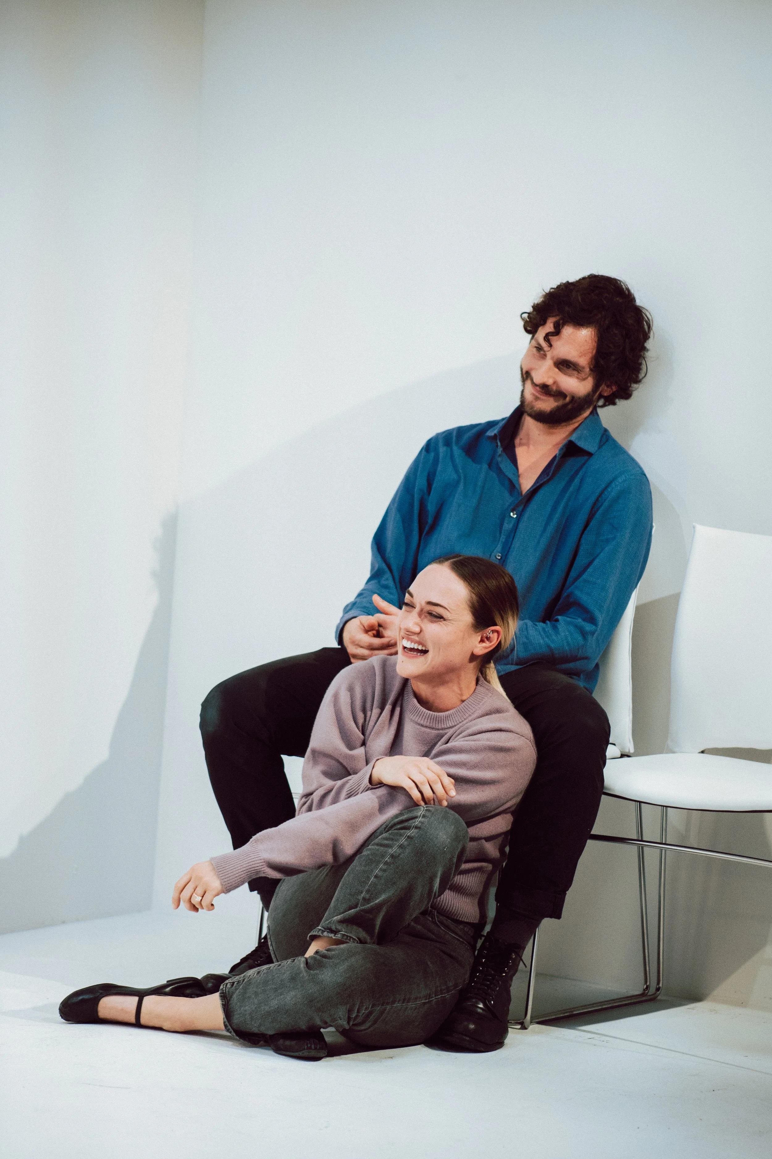 A group of three people having fun in a photography studio, with one man standing behind a woman sitting on the floor and another woman sitting on a chair, all smiling and laughing.