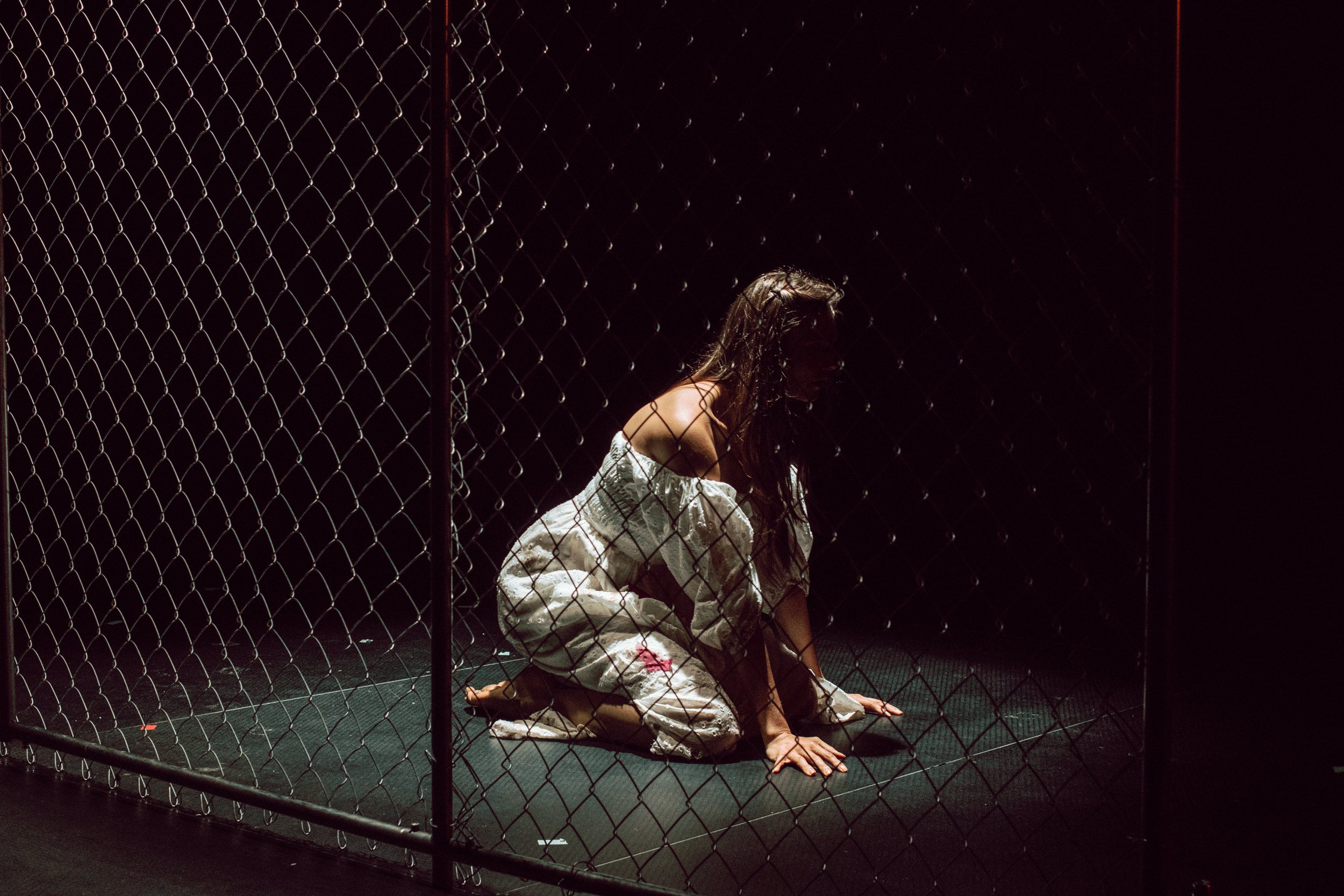 A woman with long hair, dressed in a white dress, is kneeling and sitting on her heels inside a fence enclosure. The scene is dark, with the woman illuminated by a faint light.