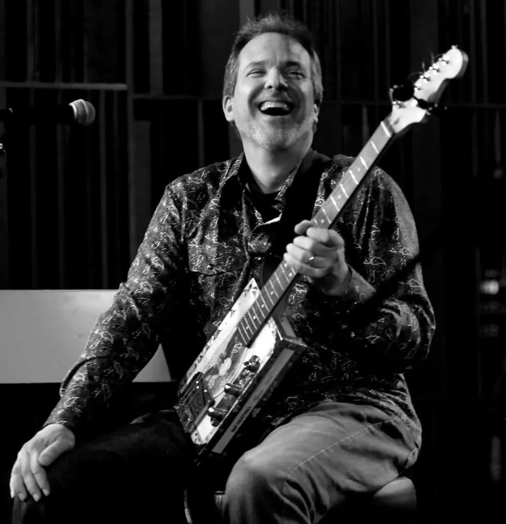 Black-and-white photo of a smiling guitarist holding a cigar box guitar.