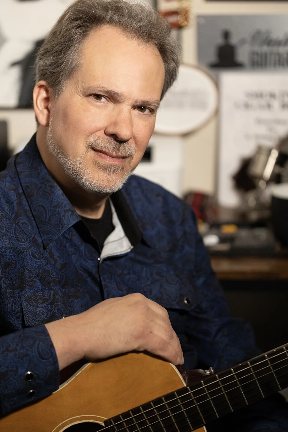 Dave Isaacs smiling, seated, wearing a blue shirt and holding an acoustic guitar.
