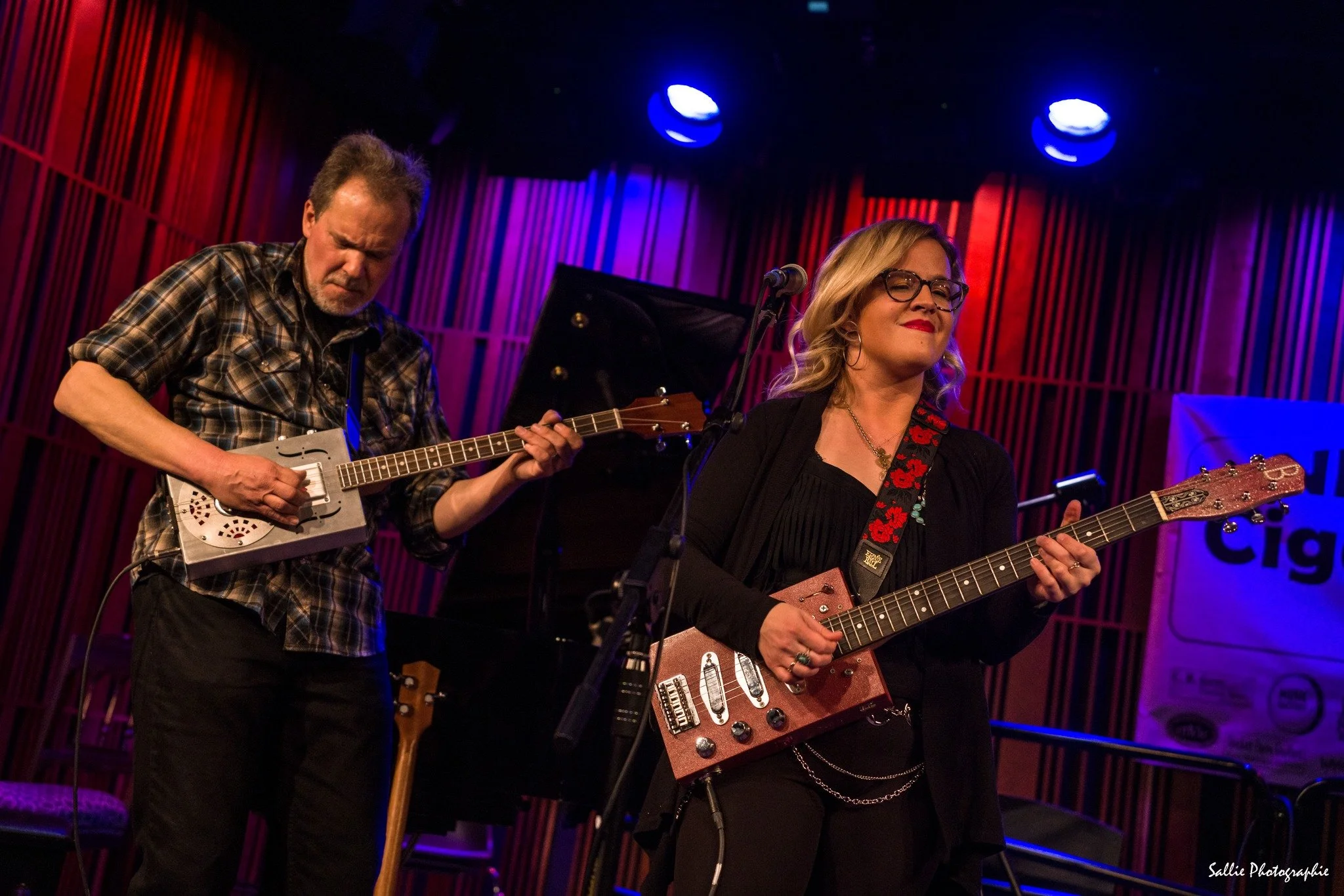 Two guitarists playing cigar box guitars onstage, a man in a plaid shirt and a woman in black.