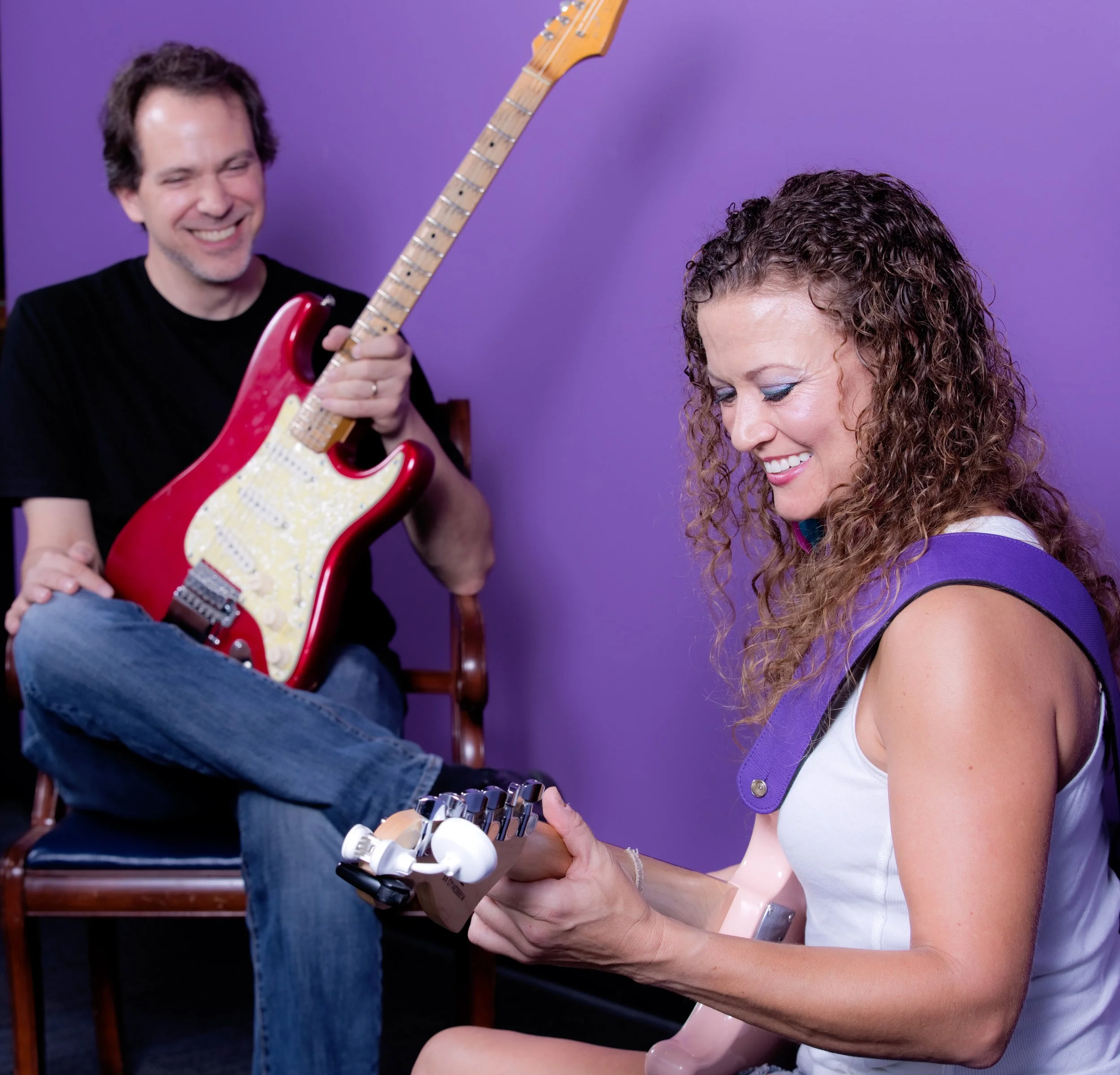 A man holding an electric guitar and a woman playing another guitar. The man is smiling and the woman is looking down with a smile. The background is purple.