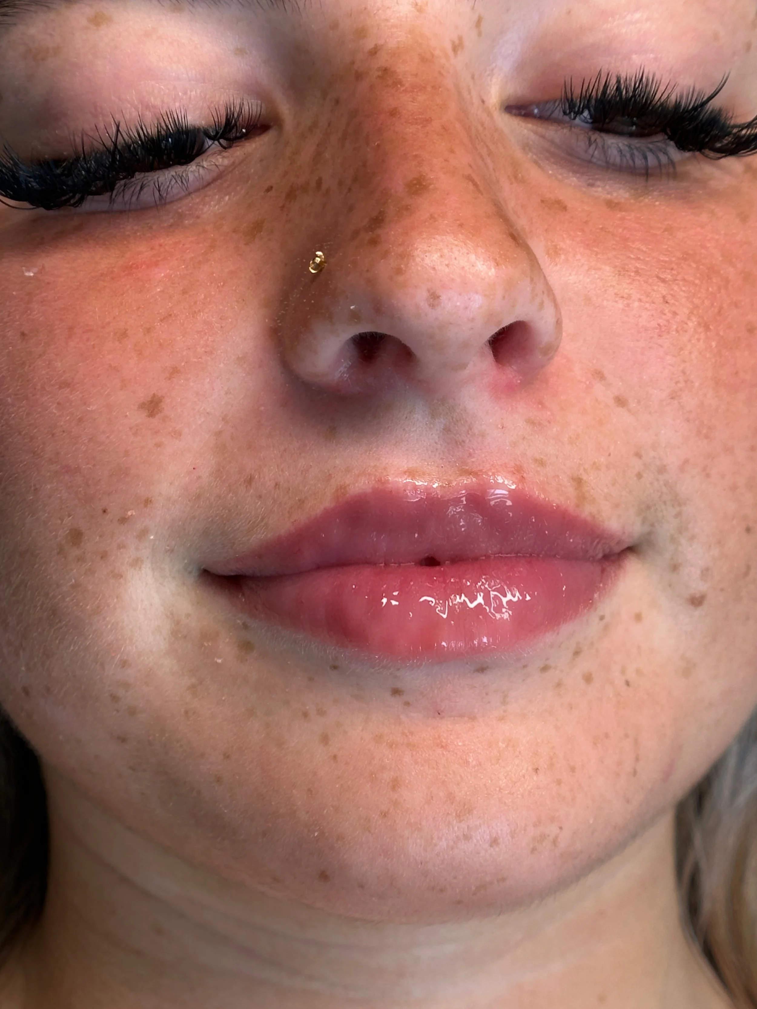 Close-up of a woman's face with freckles, closed eyes, glossy lips, and a nose piercing.