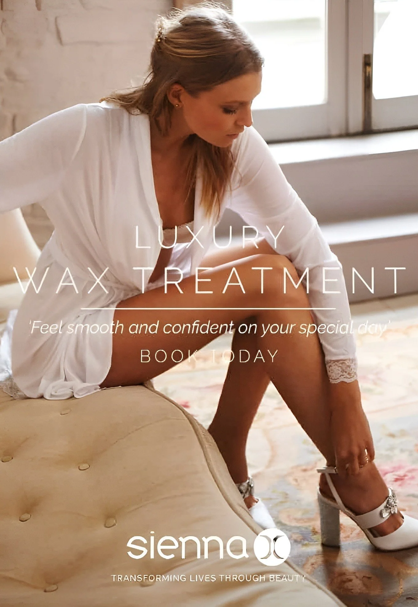 A woman in a white robe sitting on a beige ottoman, putting on a high-heeled shoe inside a bright room with large windows.