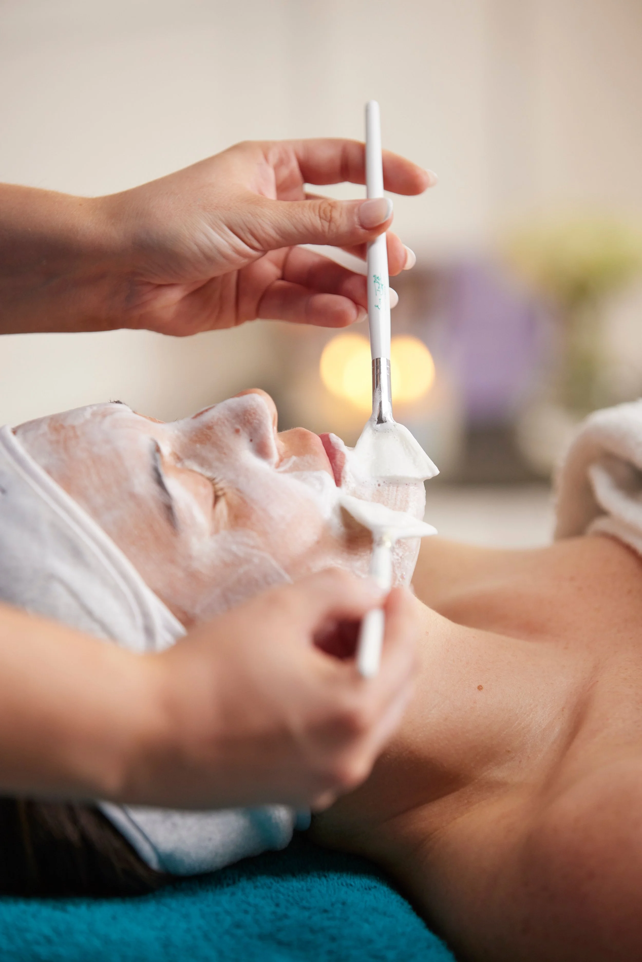 Person receiving a facial treatment with a brush, covered in a white facial mask, lying down with eyes closed.