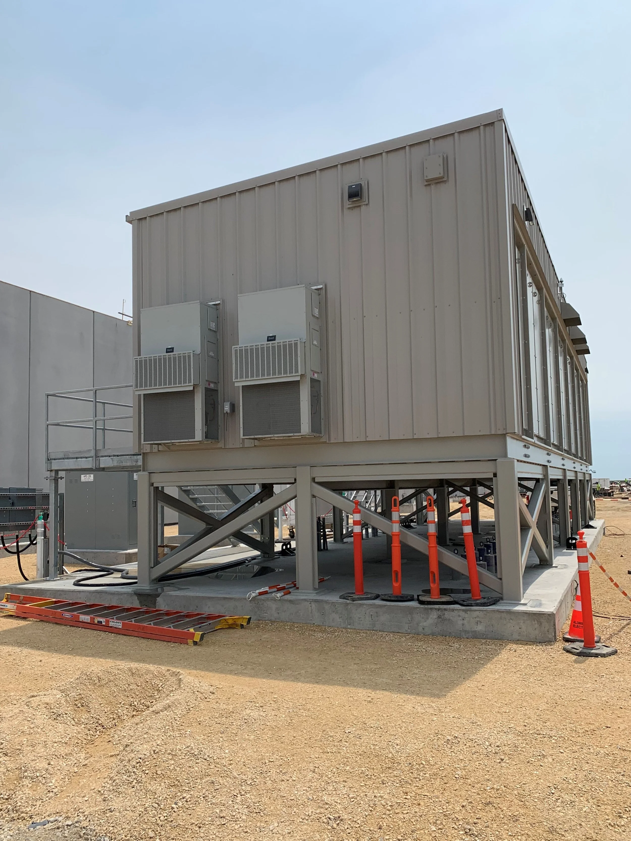 A large beige container building on a concrete platform with orange safety cones and barriers, under a clear sky.