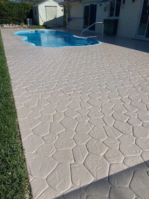 Concrete pool deck with an irregular stone pattern surrounding a blue kidney-shaped swimming pool, with a white shed or building in the background.