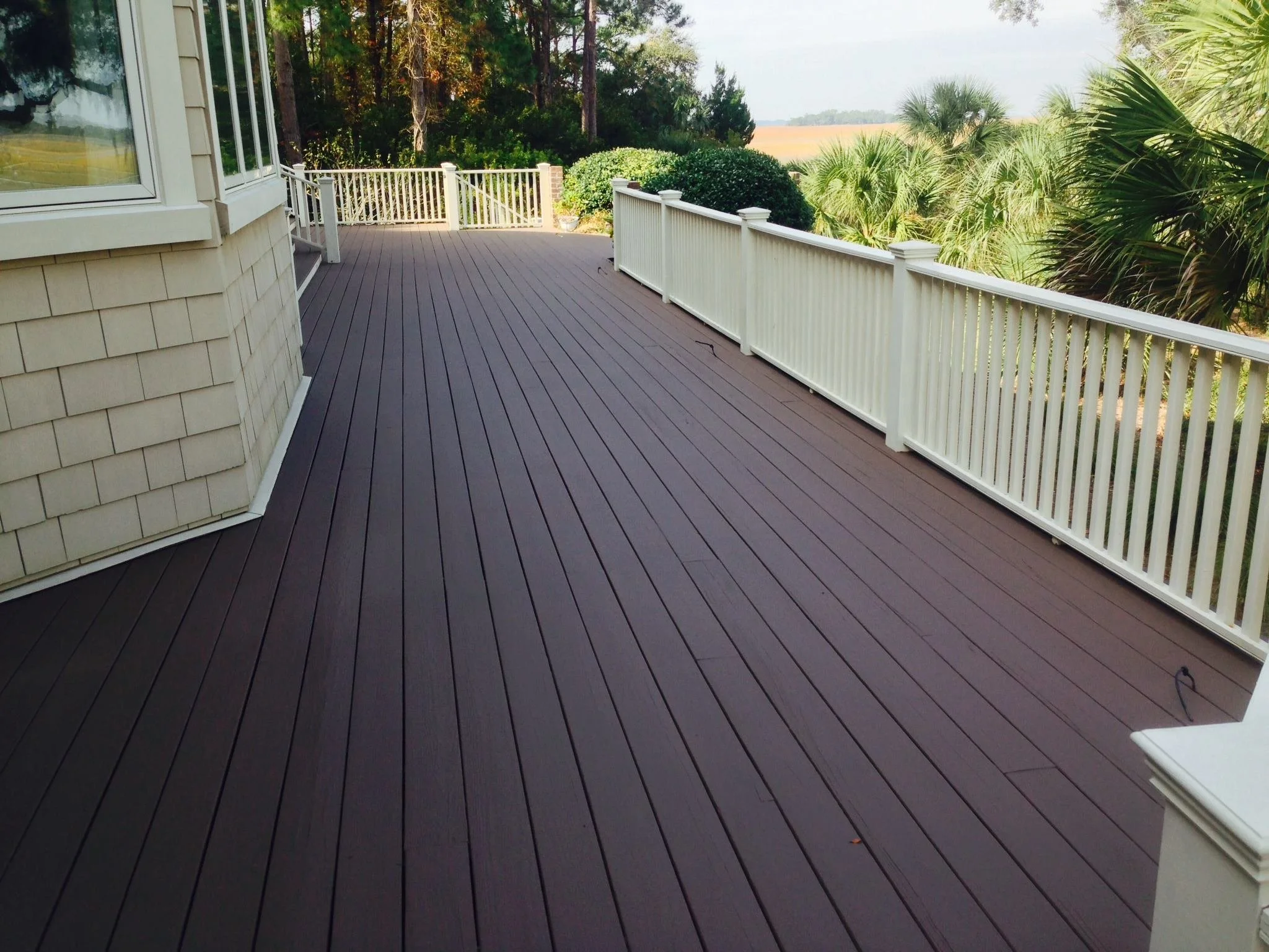 A spacious outdoor deck with dark wood flooring and white railing, attached to a house with beige siding and large windows, overlooking a lush green landscape with trees and bushes.