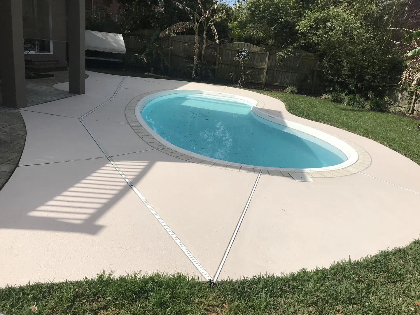 A backyard with a kidney-shaped swimming pool surrounded by a concrete deck and a grassy area, with a wooden fence and trees in the background.