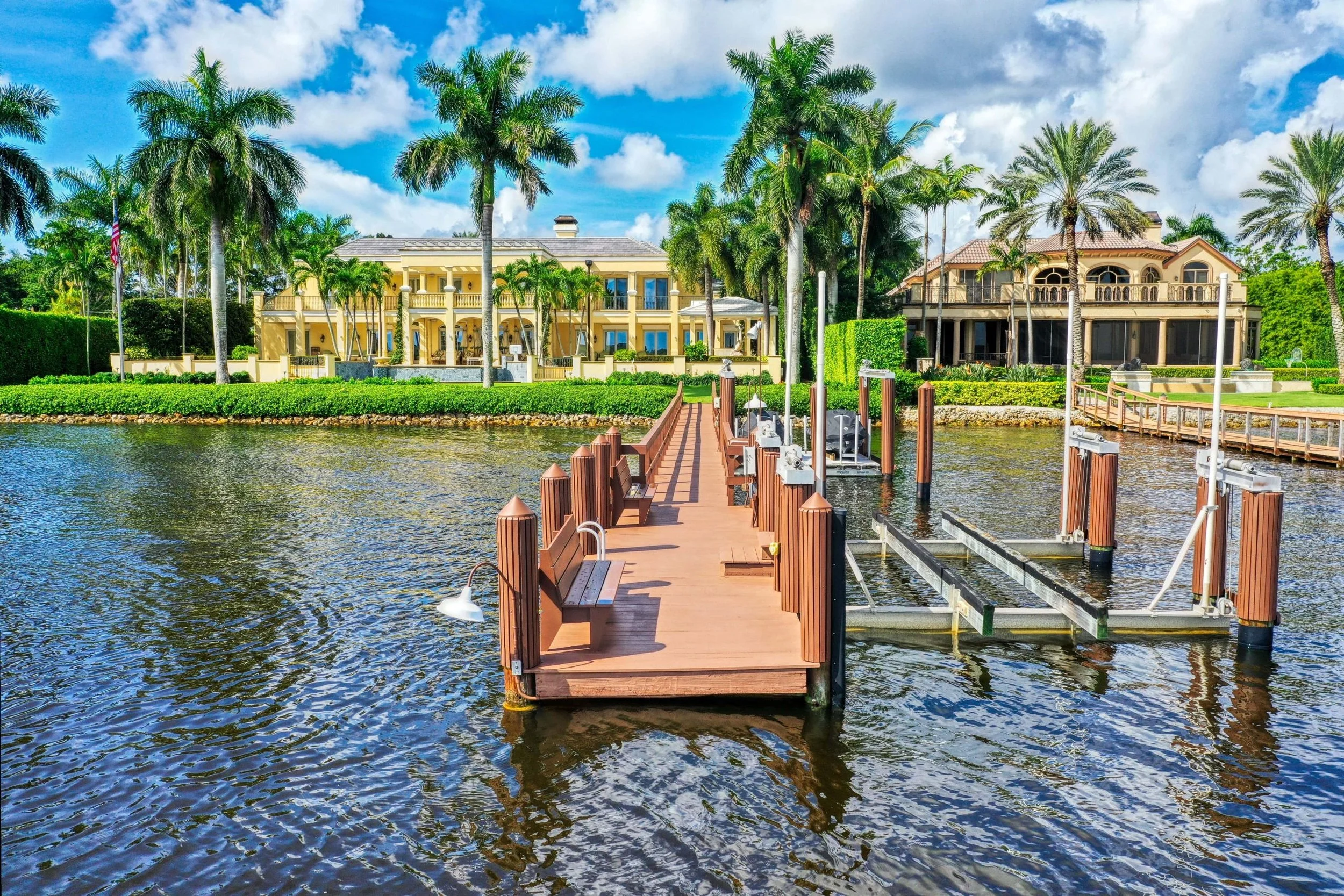 Waterfront house with palm trees, a dock with benches, and a cloudy sky.