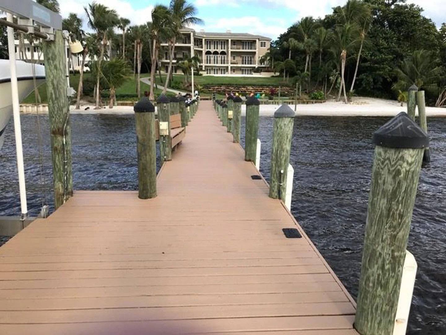 A wooden dock extending over water towards a beach with palm trees and a large waterfront building in the background.