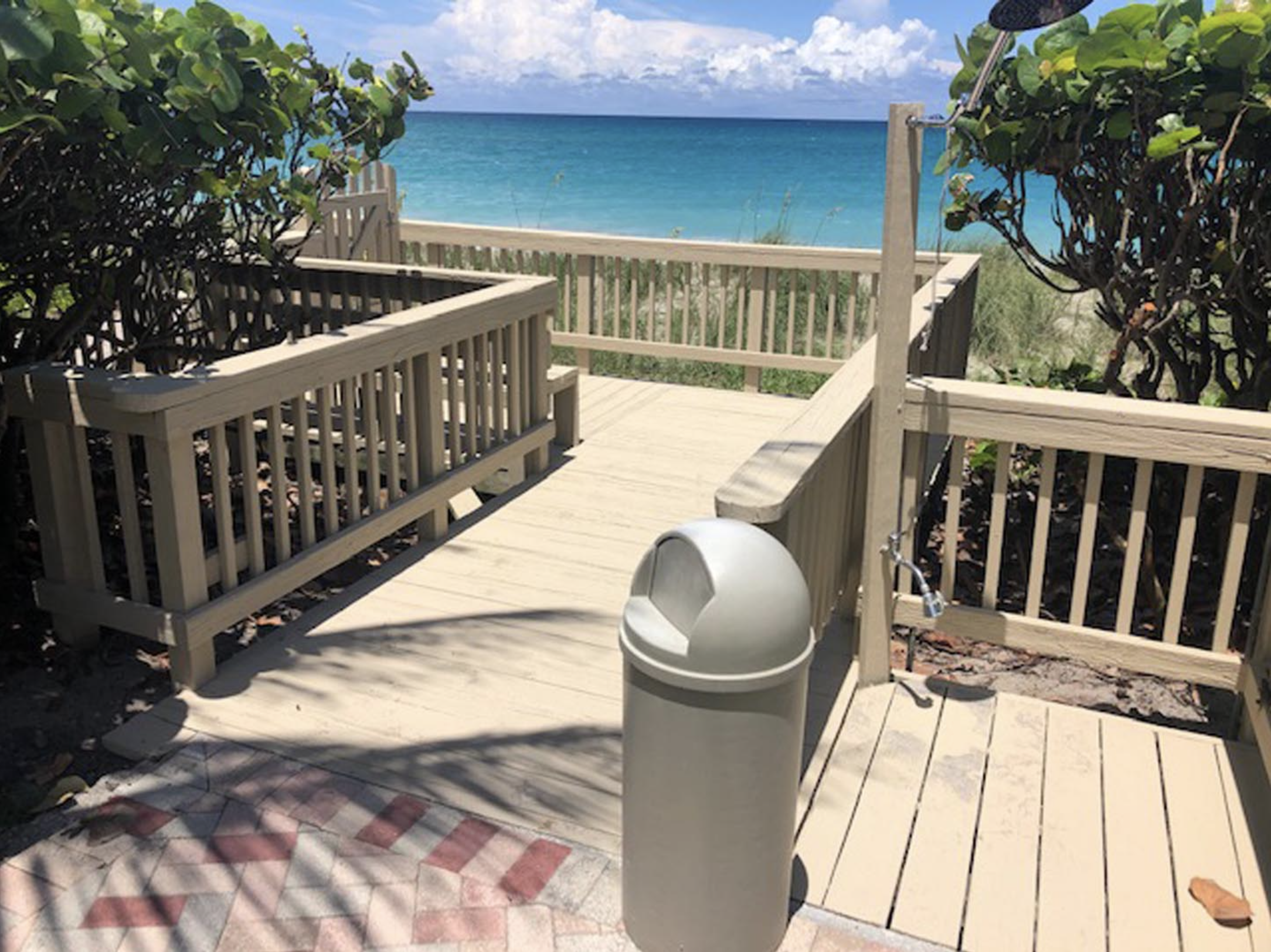 A wooden beach access deck with a railing, leading to a sandy beach and blue ocean in the background, surrounded by greenery. A trash bin is in the foreground, and a lock secures the deck gate.