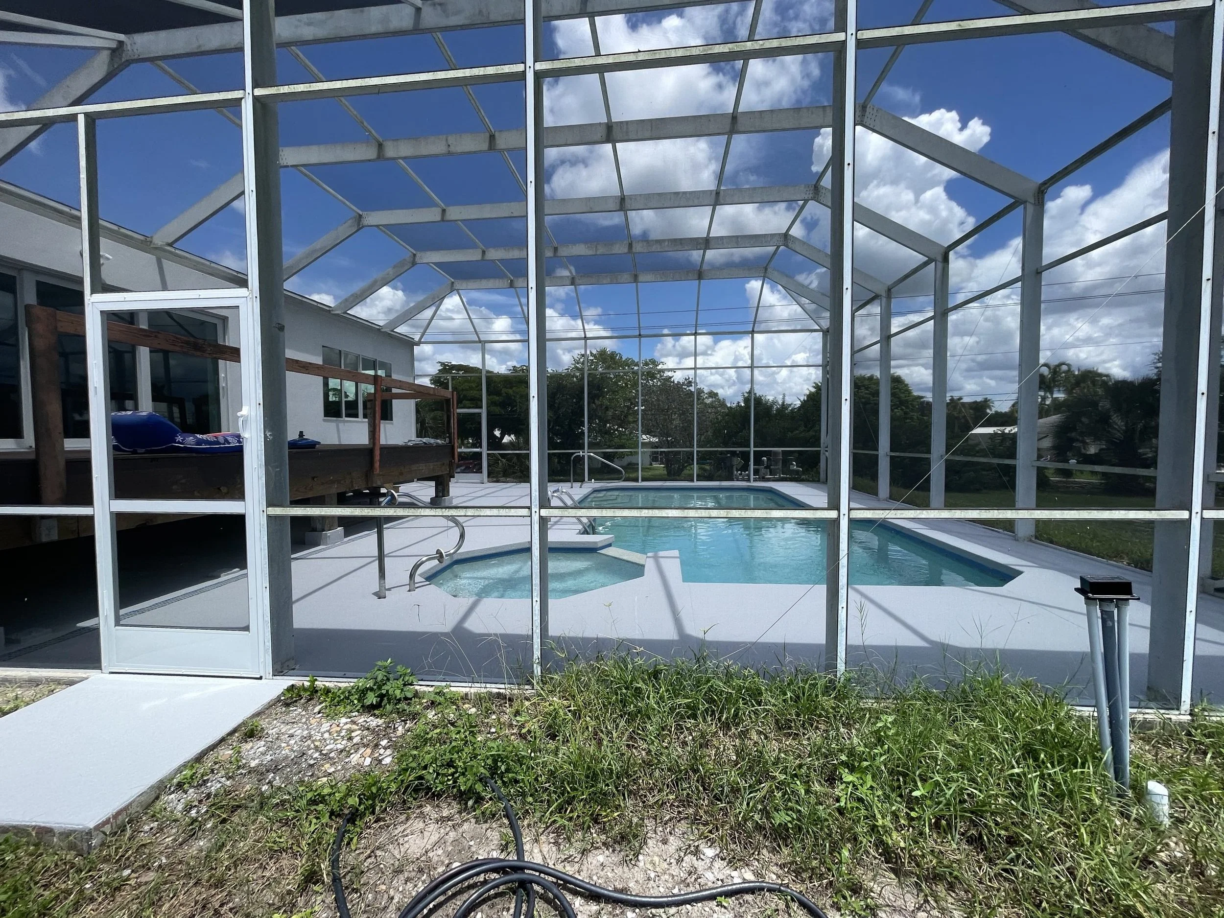 A screened-in outdoor pool and hot tub area with a clear blue sky and white clouds overhead. The pool is rectangular with a rounded hot tub connected to one end, surrounded by a concrete deck. A wooden deck is visible on the left side of the image, attached to a white building.