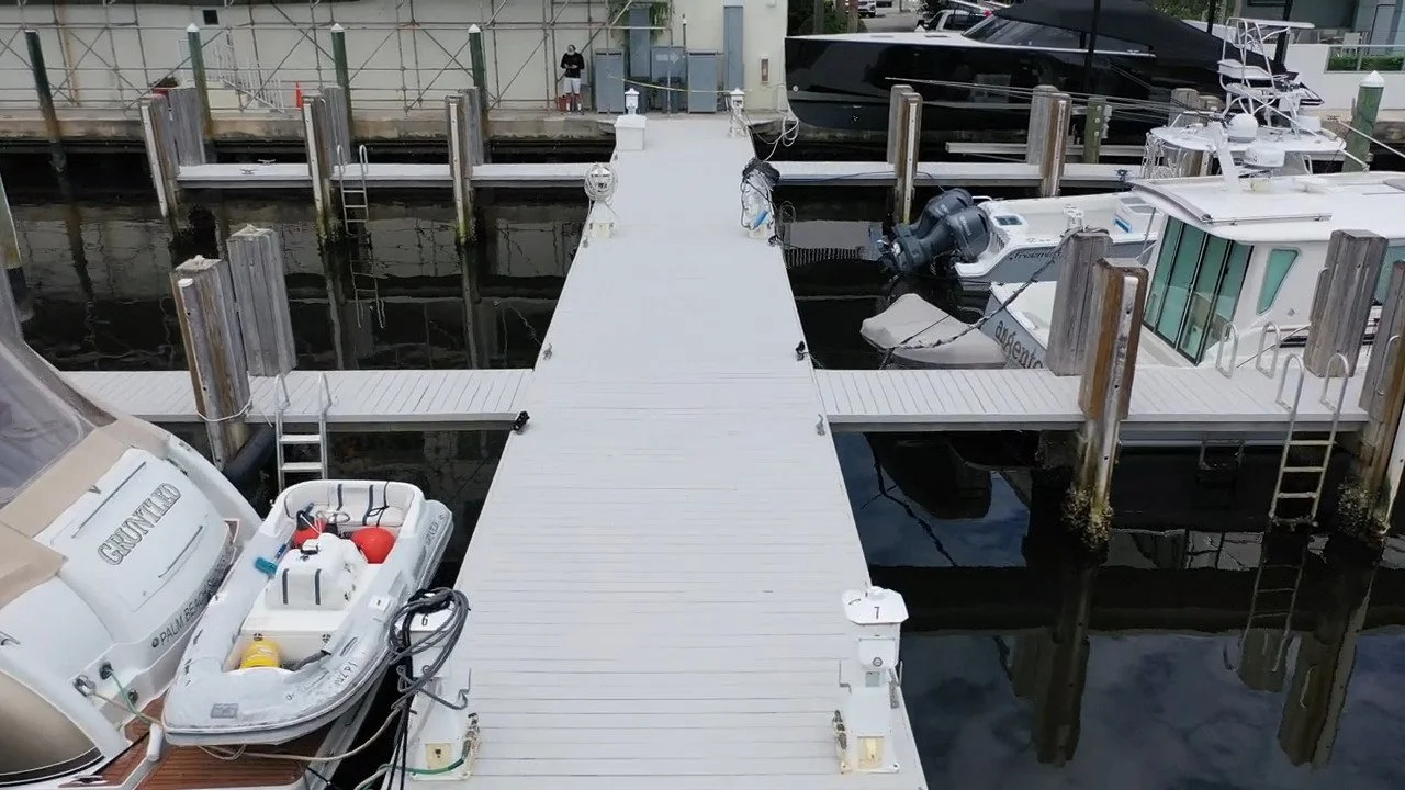A marina with a dock extending into the water, surrounded by boats including a motorboat with an outboard motor, and a black yacht in the background. There are ladders on the docks and a person standing on a boardwalk behind the deck.